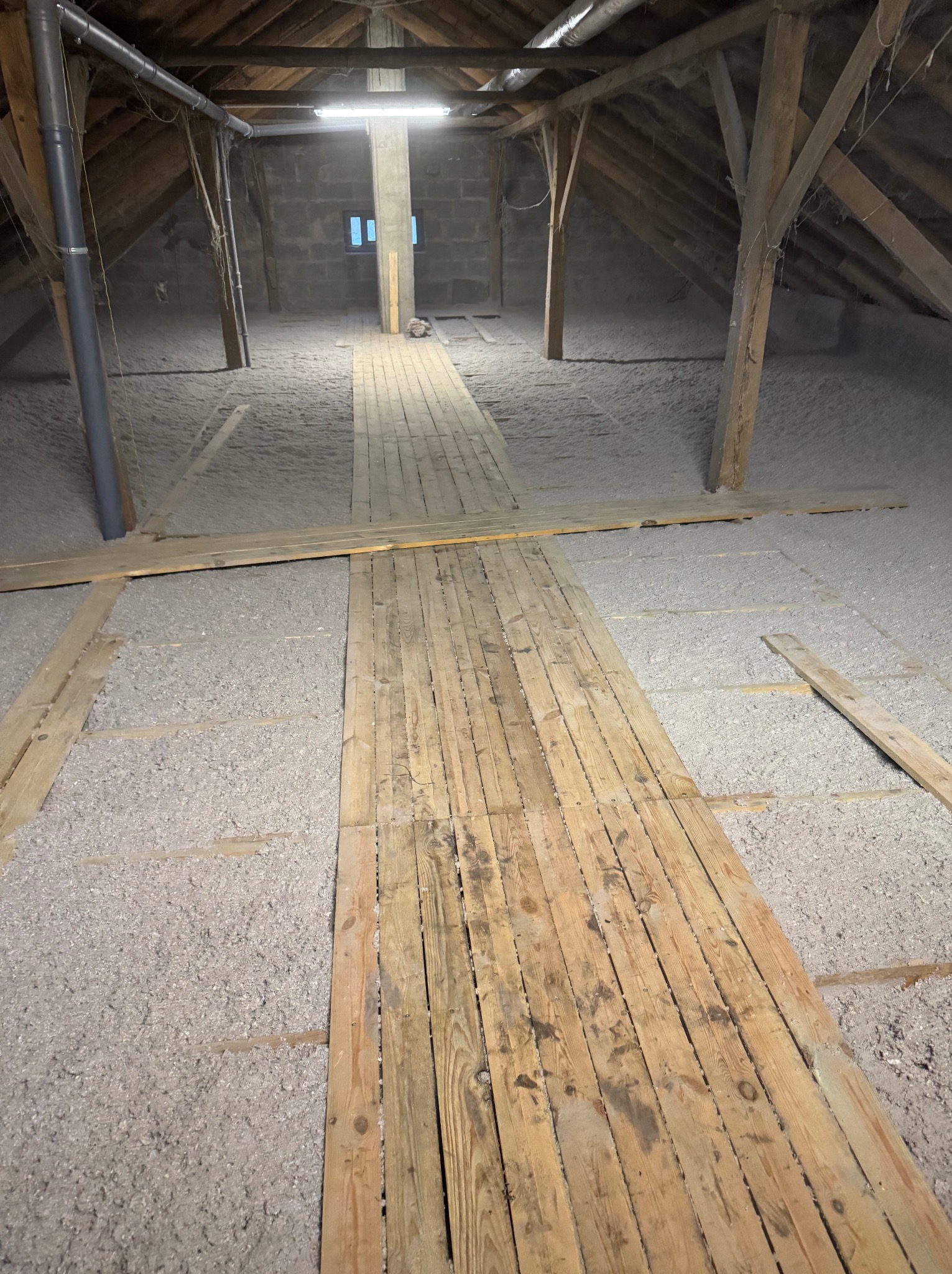 Poddasze ocieplone szarą wełną mineralną, z drewnianą kładką ułożoną wzdłuż centralnej części, widoczne drewniane krokwie i słupy konstrukcyjne, oraz fragment instalacji wentylacyjnej z szarej rury...