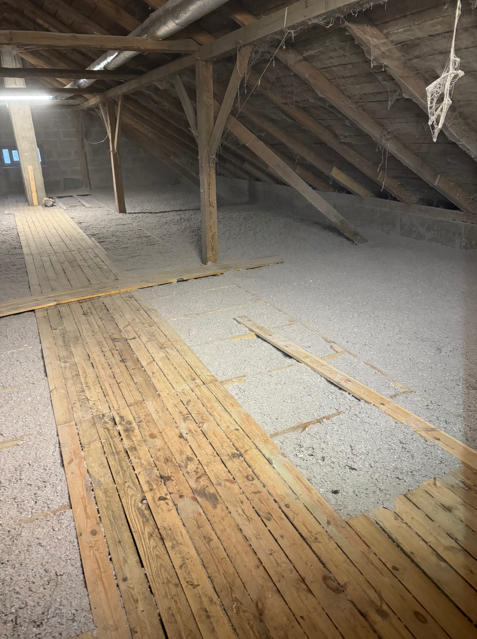 Poddasze z drewnianą konstrukcją i częściowo ułożoną podłogą z desek, pokryte szarą warstwą izolacyjną z wełny mineralnej, widoczne pajęczyny i rury wentylacyjne pod sufitem.