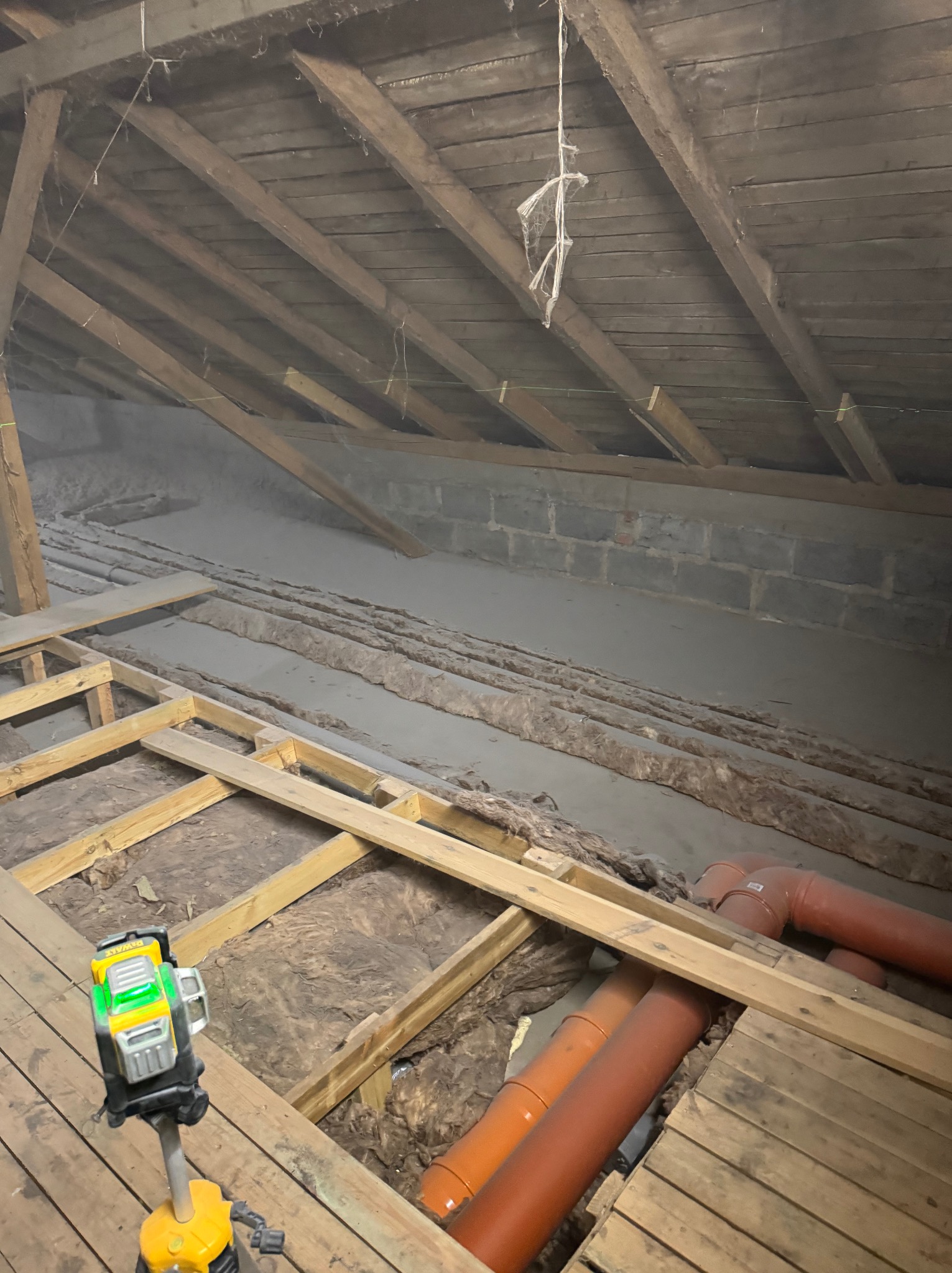 Poddasze z widoczną izolacją z wełny mineralnej między drewnianymi belkami stropowymi, częściowo pokryte deskami podłogowymi, z widocznymi rurami wentylacyjnymi i poziomnicą laserową.