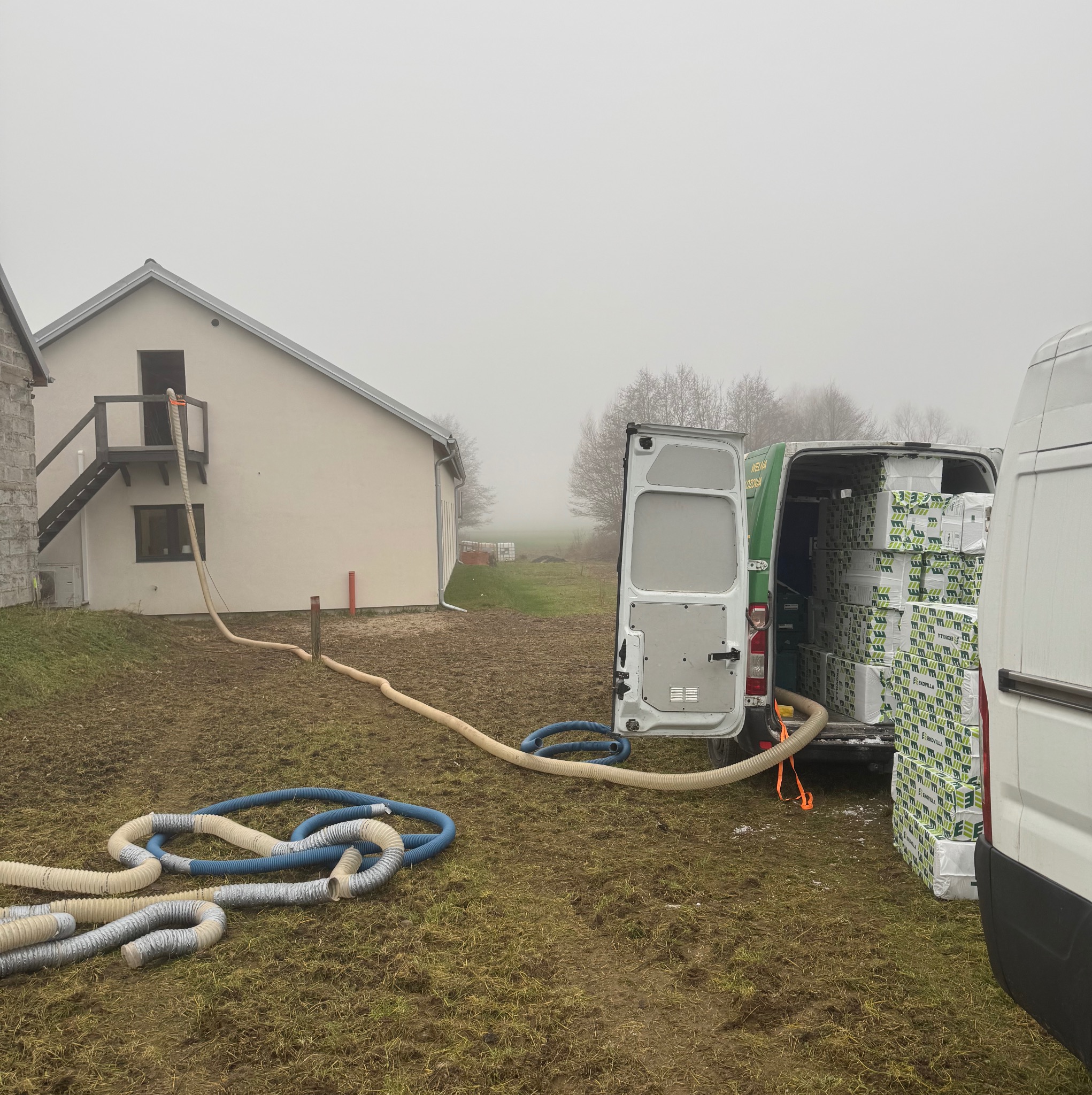 Ocieplanie poddasza wełną celulozową: biały wąż prowadzi z samochodu dostawczego wypełnionego paczkami wełny do otworu w ścianie domu, na zewnątrz pochmurny dzień.