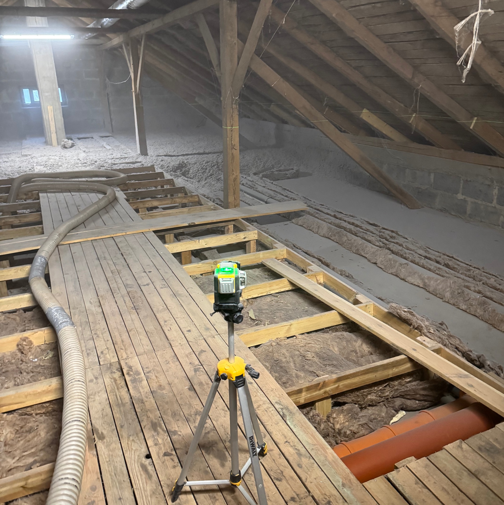 Poddasze w trakcie ocieplania wełną mineralną, widoczny laser krzyżowy na statywie i rura do podawania materiału izolacyjnego.