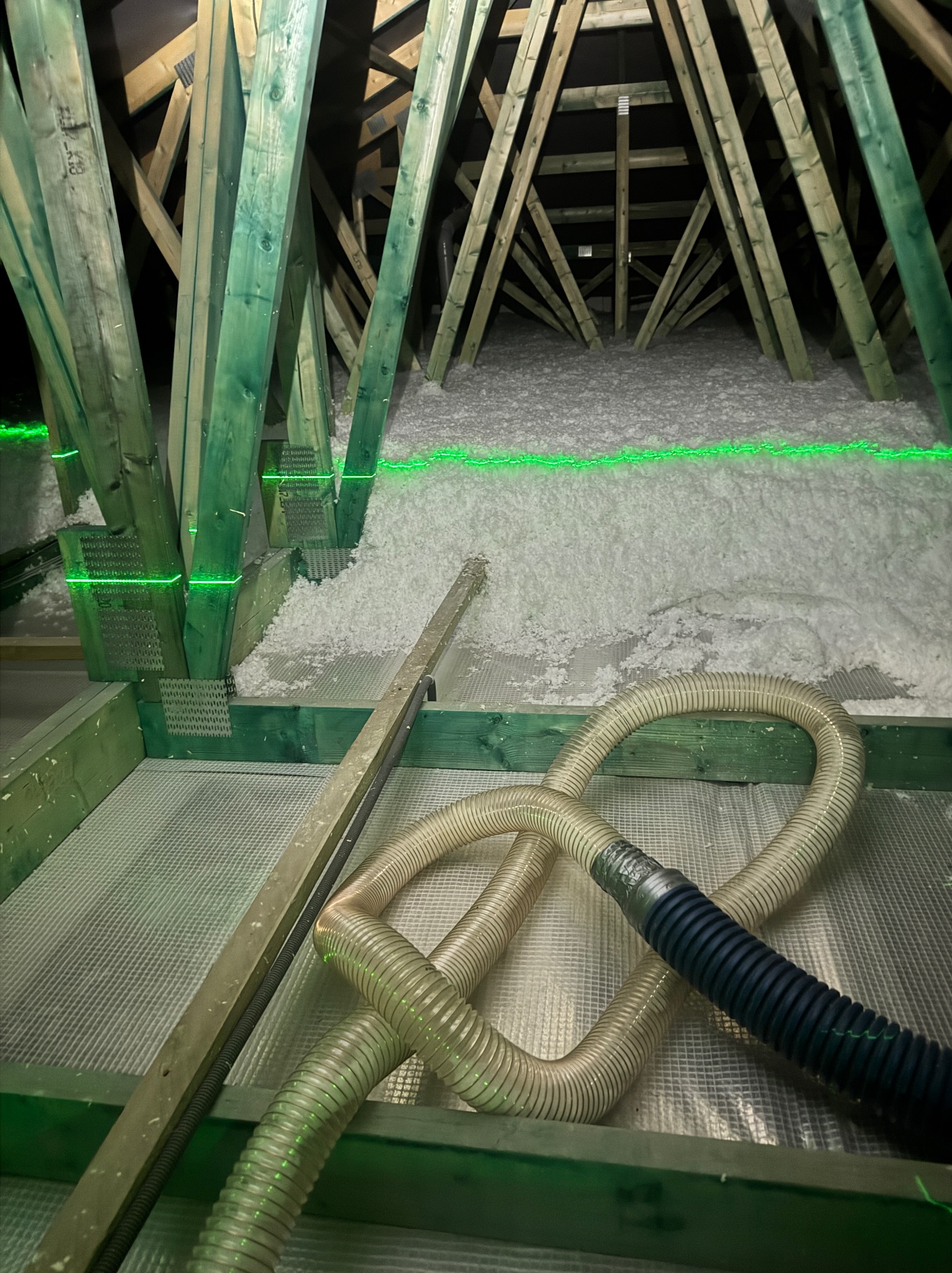 Poddasze z drewnianą konstrukcją, wypełnione białą izolacją. Widoczny wąż do aplikacji izolacji i zielona linia lasera wyznaczająca poziom.