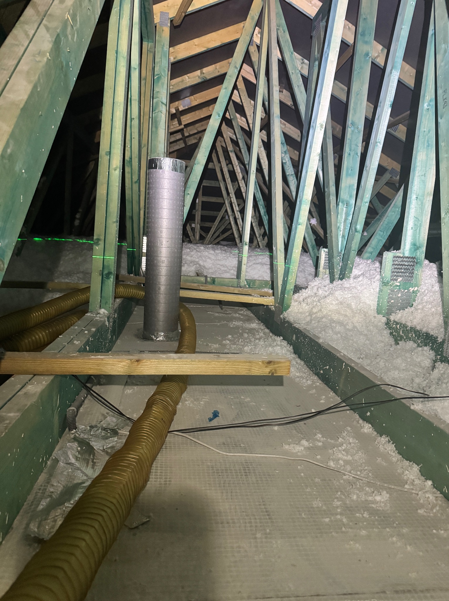 Poddasze z drewnianą konstrukcją, ocieplone białą wełną mineralną. Widoczne rury wentylacyjne i zielony laser poziomujący. Prace ociepleniowe w toku.