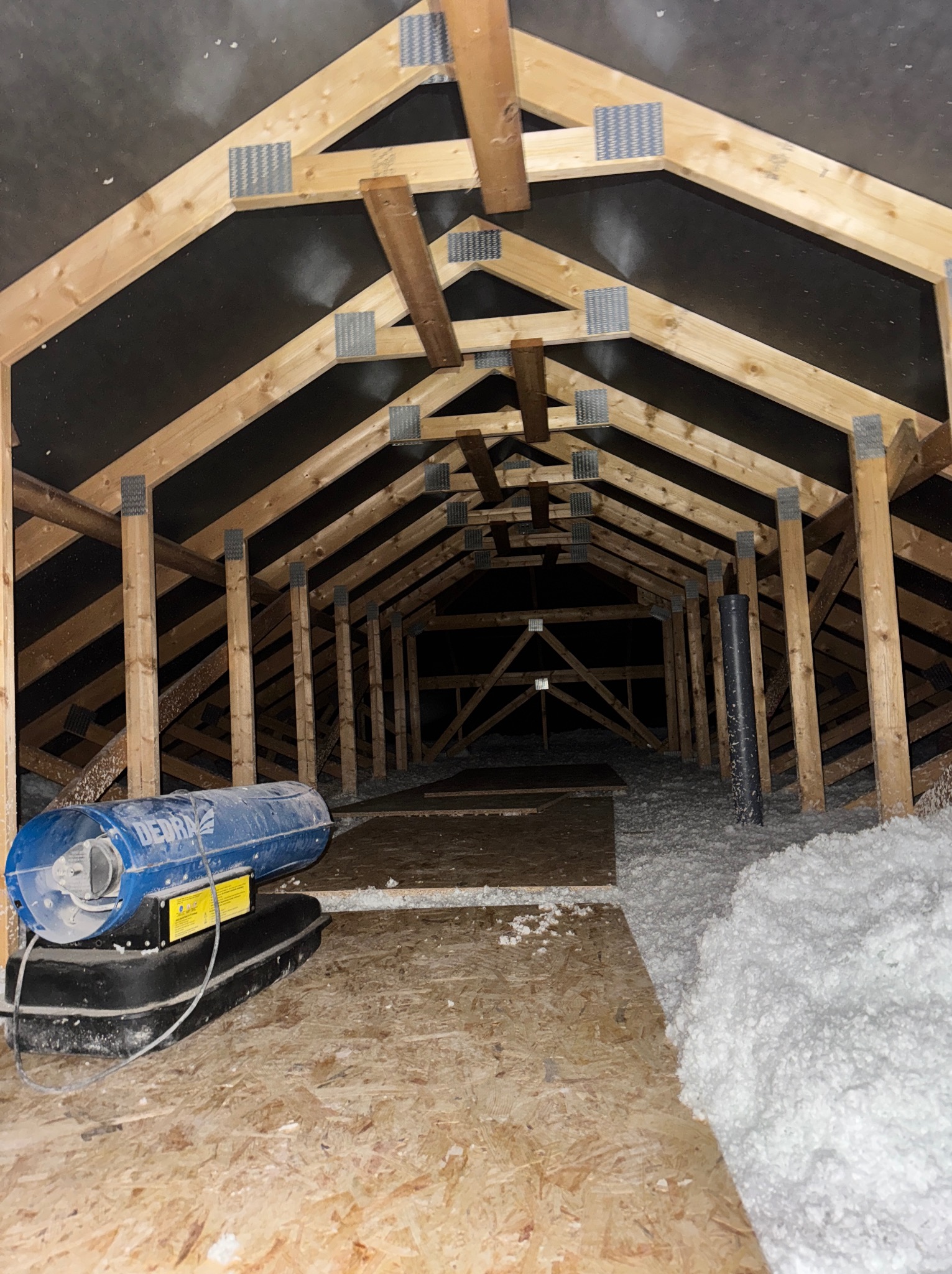 Poddasze z drewnianą konstrukcją więźby dachowej, częściowo ocieplone białą wełną mineralną. Widoczny agregat do wdmuchiwania izolacji. Ujęcie z dołu, w głąb poddasza.