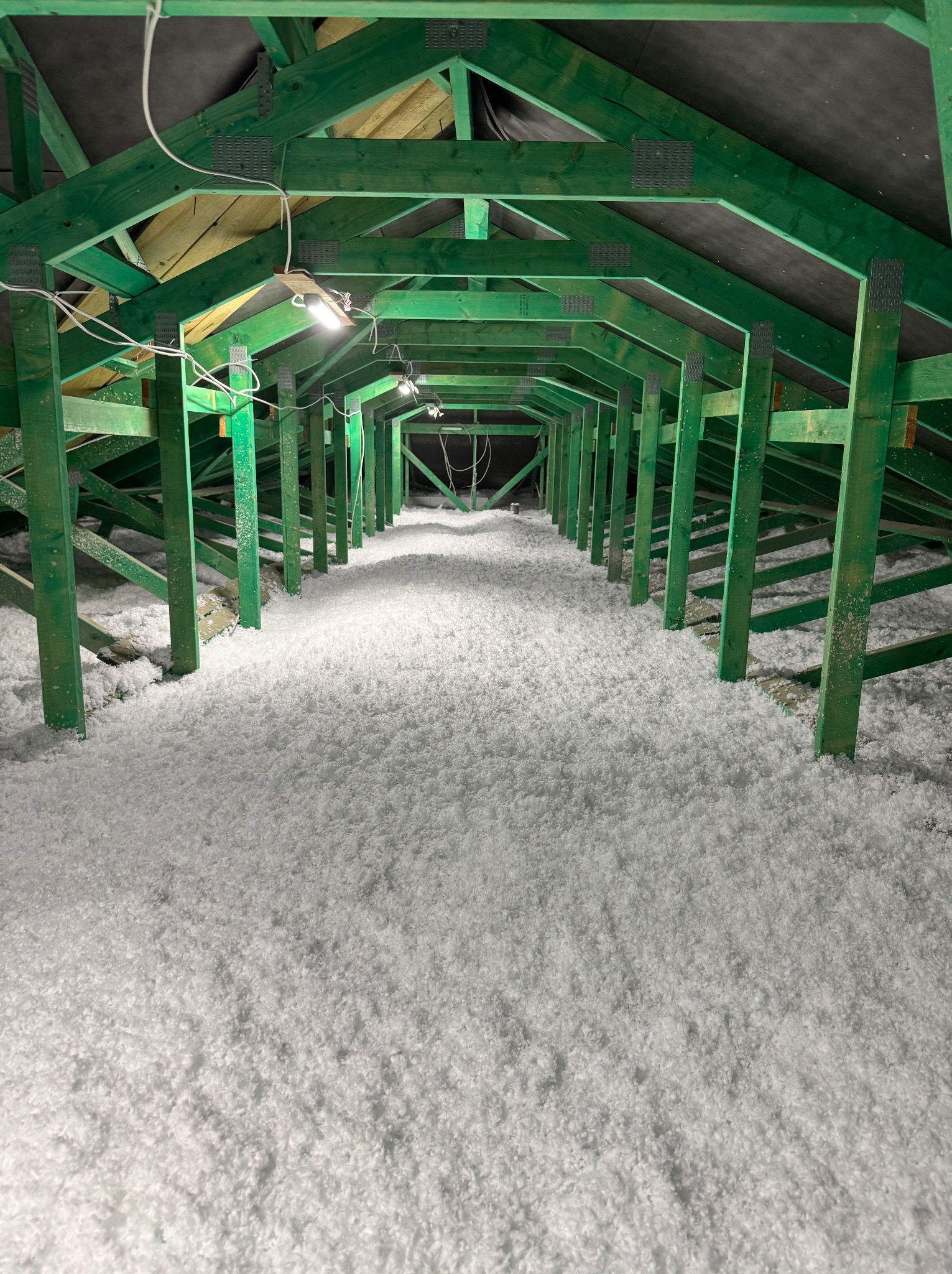 Poddasze po izolacji w Toruniu. Widok na zieloną konstrukcję dachu wypełnioną białą warstwą izolacyjną. Oświetlenie robocze. Perspektywa z głębi poddasza.