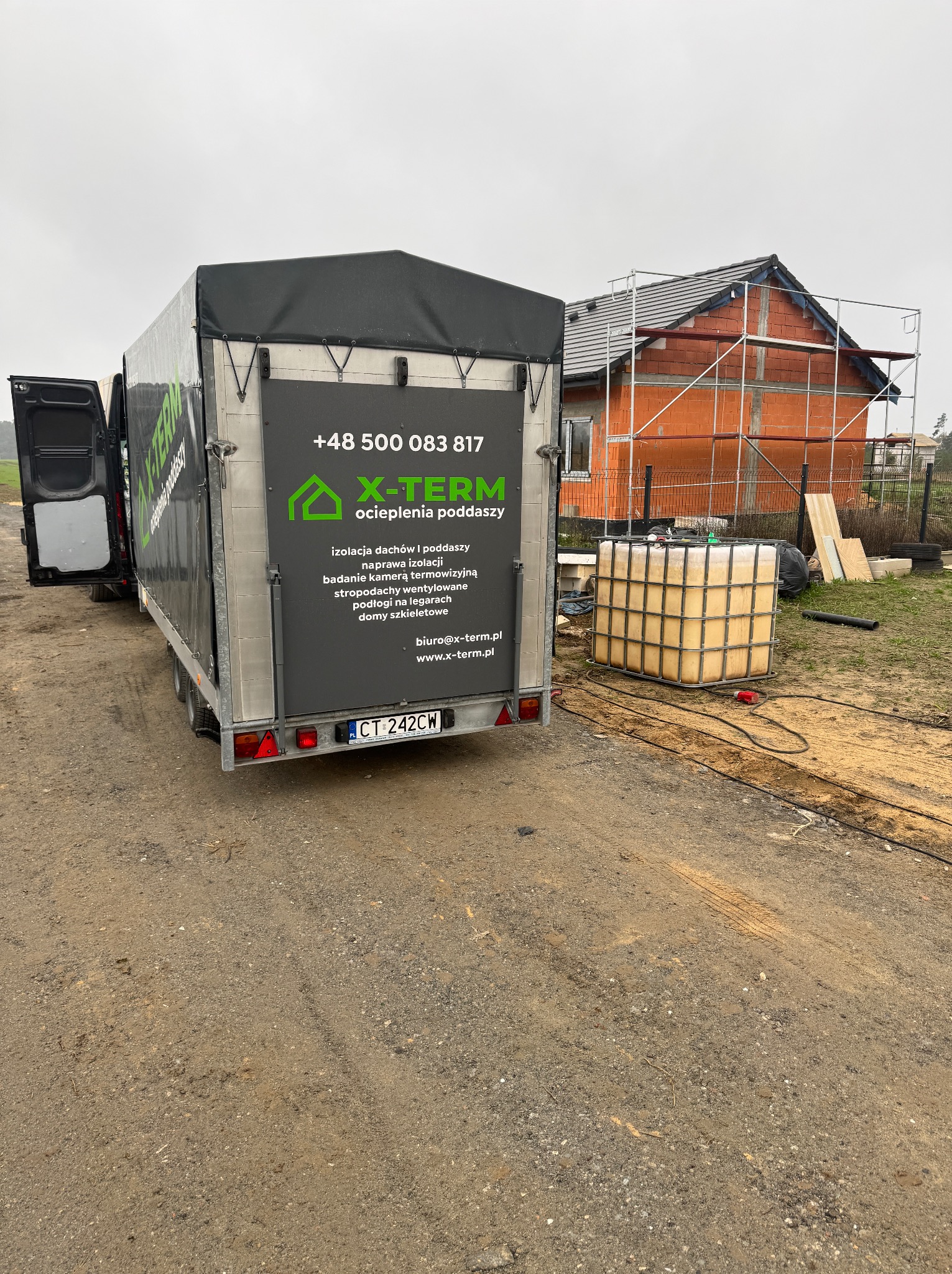 Przyczepa firmy X-Term z reklamą ocieplenia poddaszy na tle budowy domu w stanie surowym otwartym. Widoczny zbiornik z materiałem izolacyjnym.
