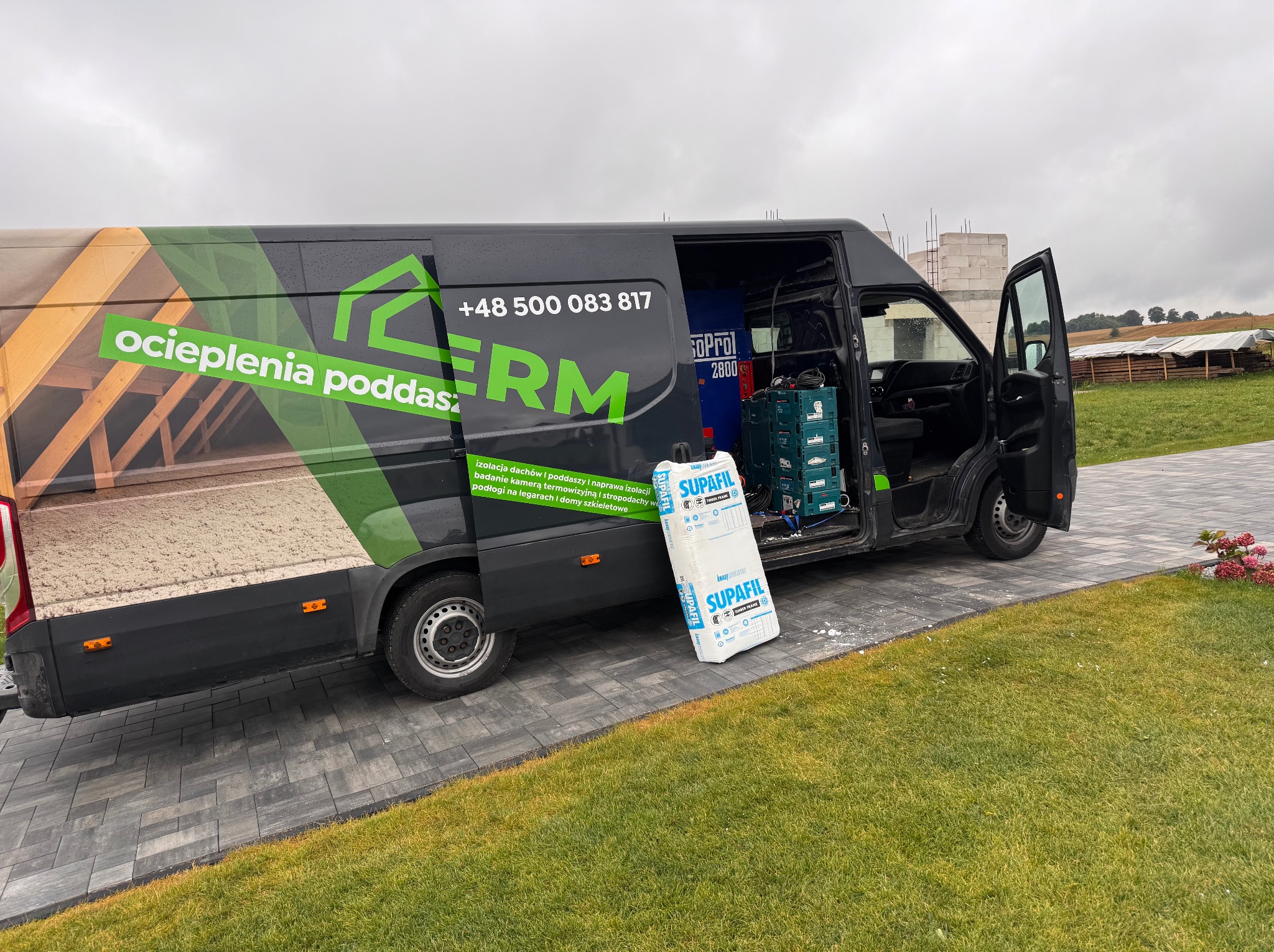 Czarny van z grafiką ocieplenia poddasza i logo firmy, otwarte drzwi boczne, wewnątrz sprzęt. Obok paczka izolacji Supafil. W tle budowa i zielony krajobraz.