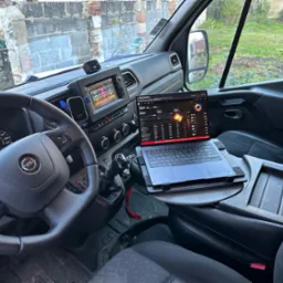 Wnętrze samochodu dostawczego z laptopem na specjalnym stoliku, wyświetlającym wykresy giełdowe, z widokiem na ceglany budynek przez przednią szybę.