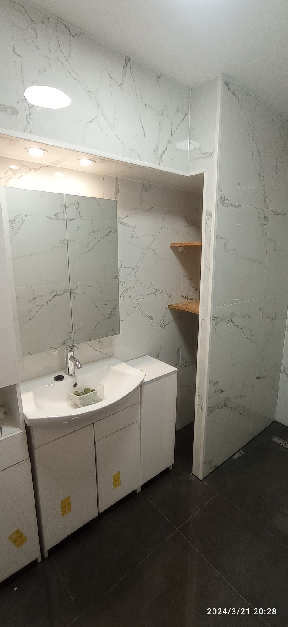 Wykończona łazienka z umywalką nablatową, szafką pod umywalką i lustrem, wykończona płytkami imitującymi marmur z półkami w niszy. Na blacie umywalki mała roślina.