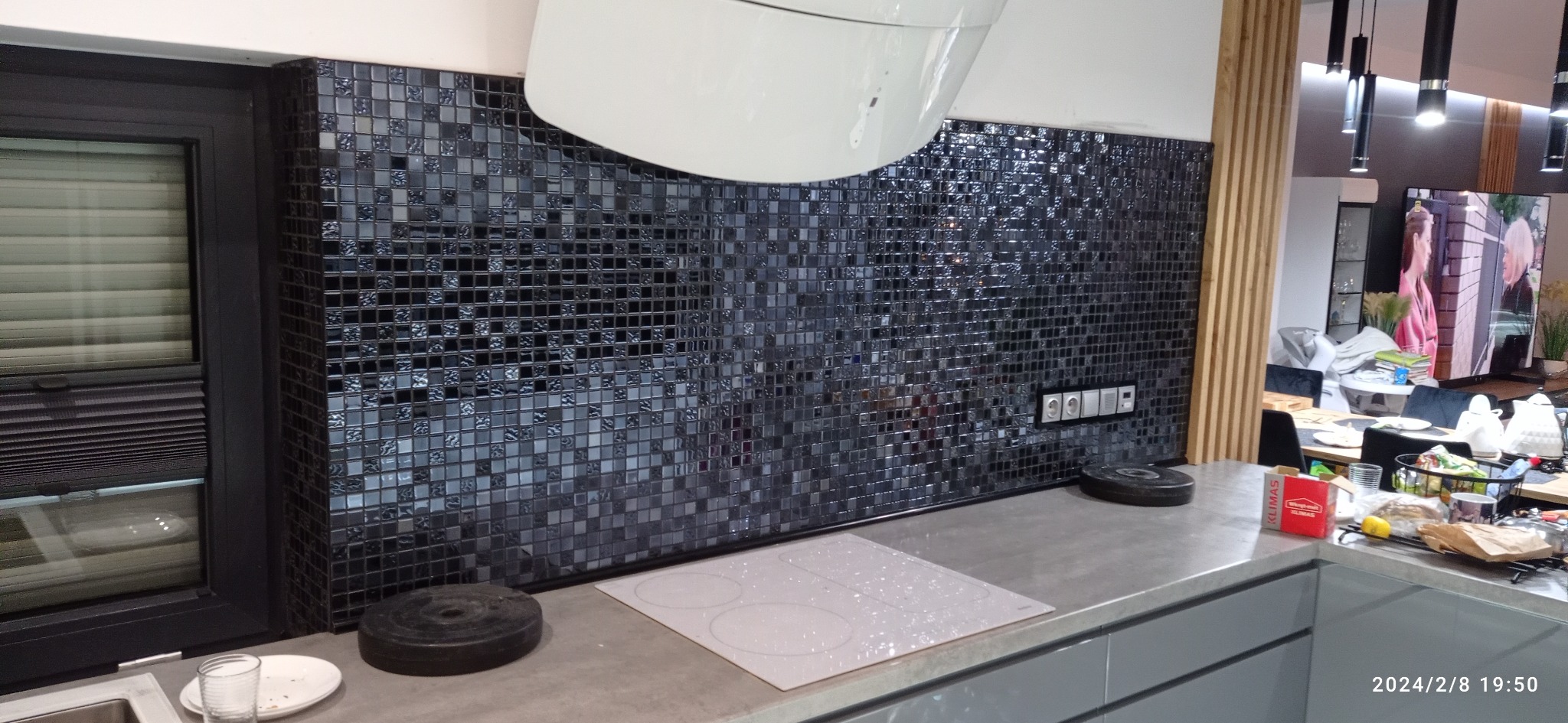 Błyszcząca mozaika z drobnych, czarnych płytek na ścianie w kuchni, nowoczesna biała płyta indukcyjna, szary blat, widoczne gniazdka i włączniki, okno z żaluzjami w tle.