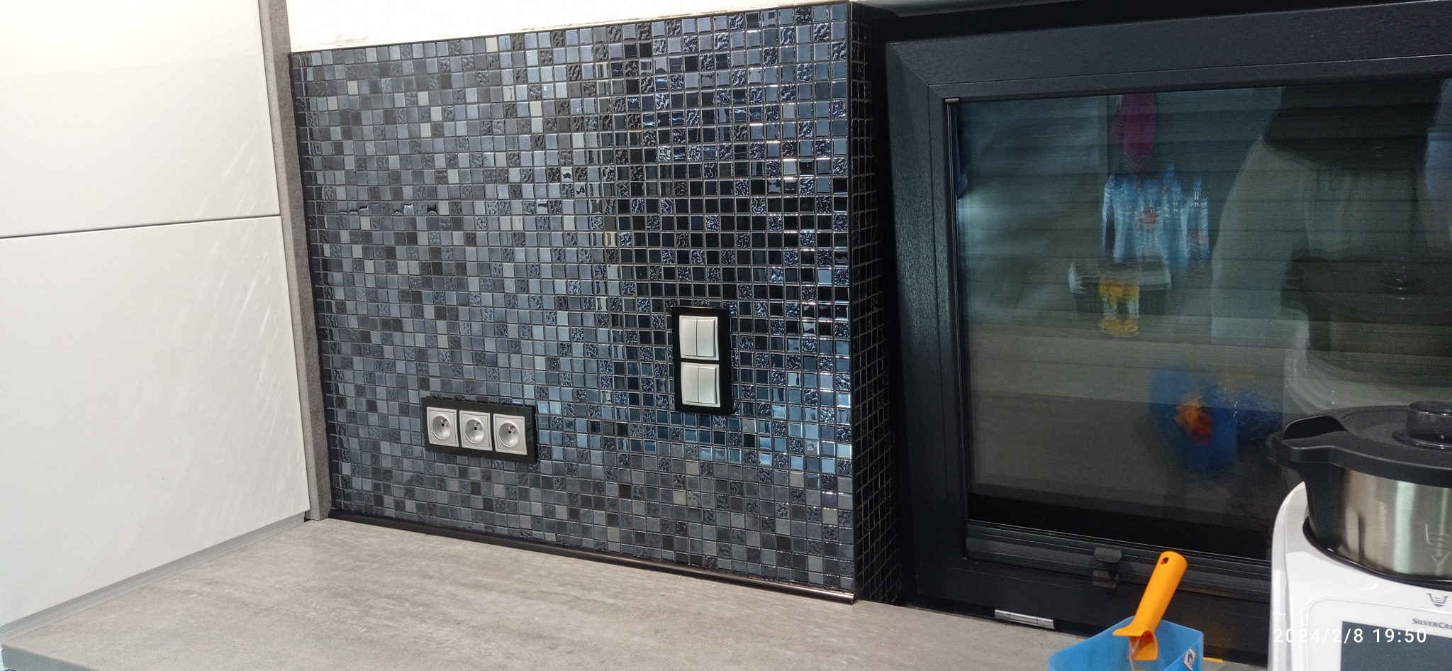 Mozaika z ciemnych, błyszczących płytek w kuchni, z widocznymi gniazdkami elektrycznymi i włącznikiem światła, sąsiadująca z czarnym oknem.