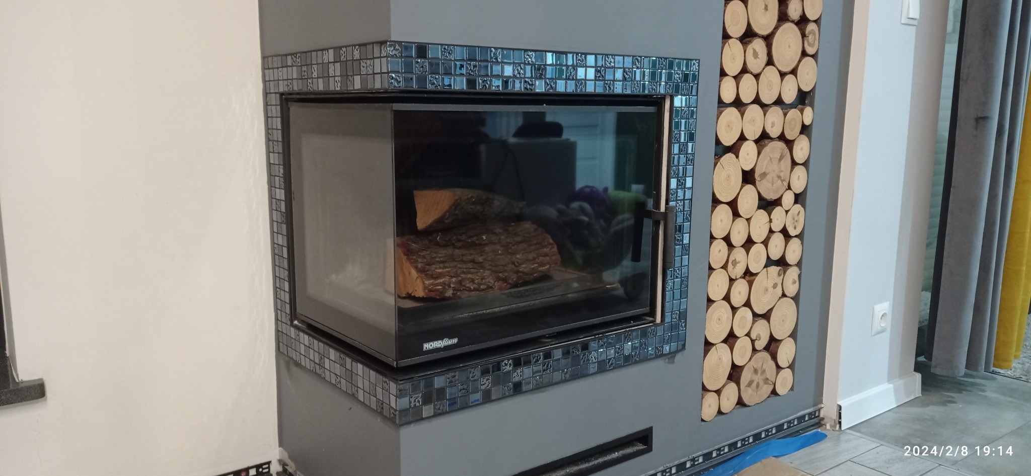 Nowoczesny kominek z szybą i dekoracyjną obudową z mozaiki, obok ściana zdobiona plastrami drewna