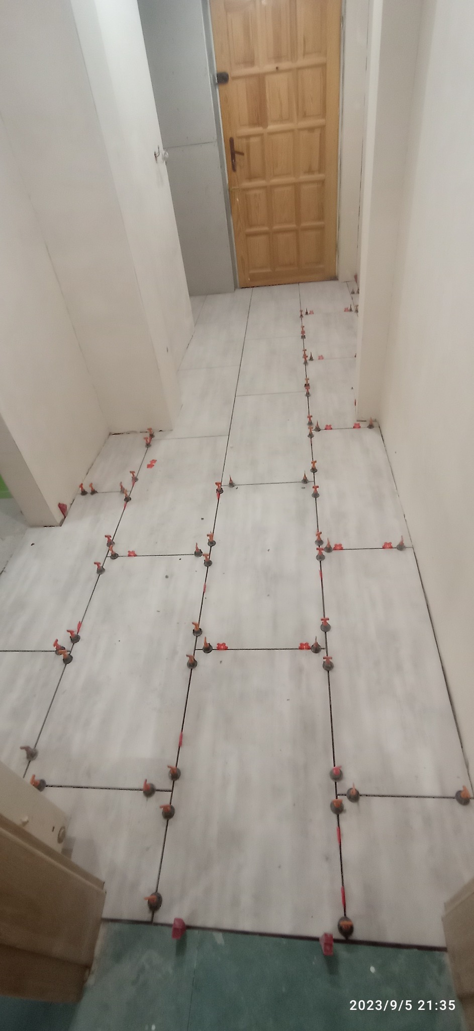 Ułożone szare płytki podłogowe w korytarzu, z widocznymi systemami poziomowania płytek w kolorze czerwonym i czarnym, drewniane drzwi w tle, ściany przygotowane pod malowanie.