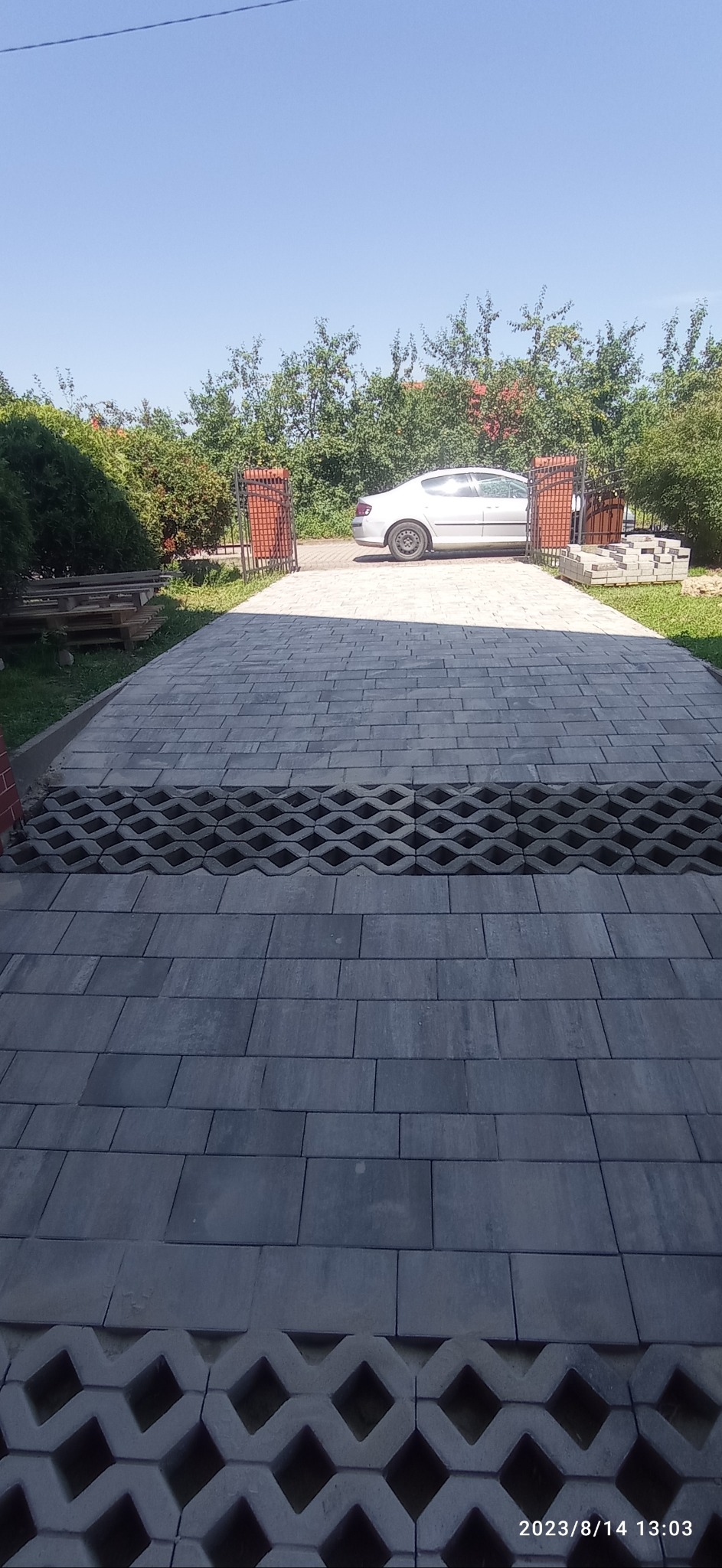 Ułożona szara kostka brukowa na podjeździe, z elementami ażurowymi, w tle otwierana brama i zaparkowany srebrny samochód.