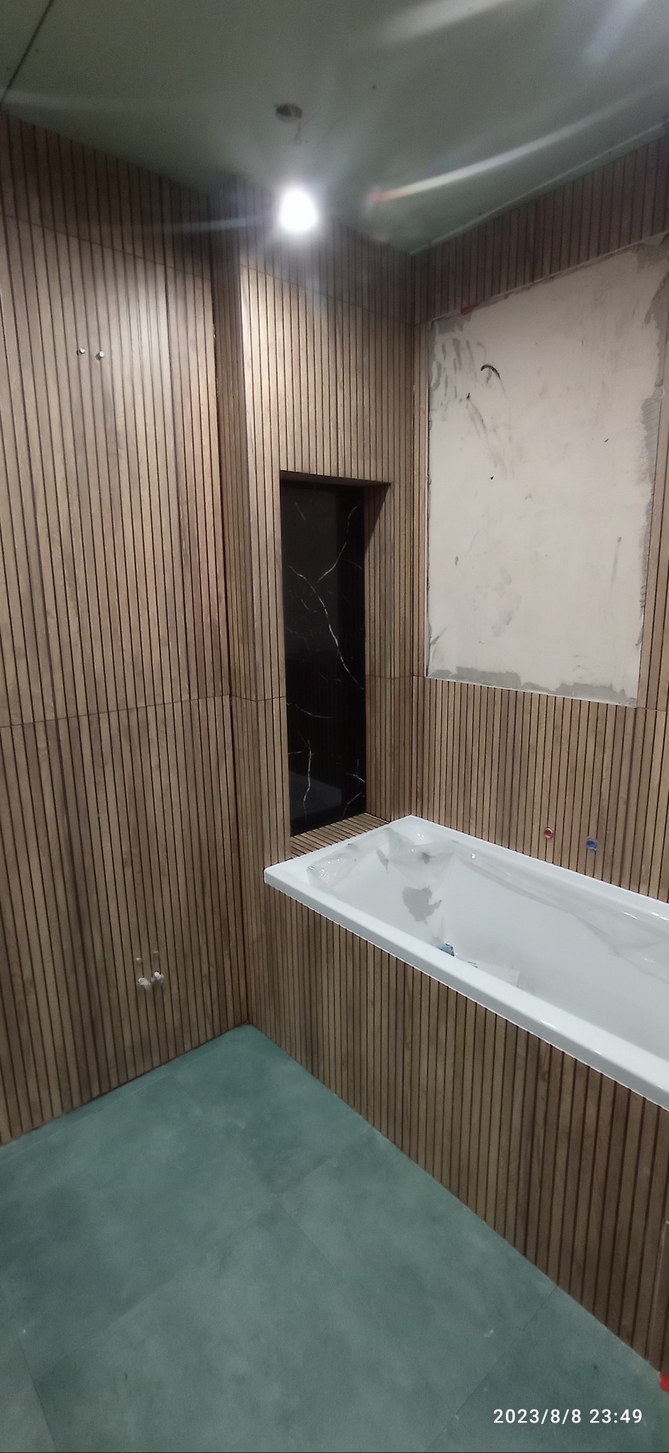 Łazienka w trakcie remontu: drewniane panele ścienne, biała wanna w folii ochronnej, zielona podłoga, czarna wnęka, fragment ściany przygotowany do położenia płytek.