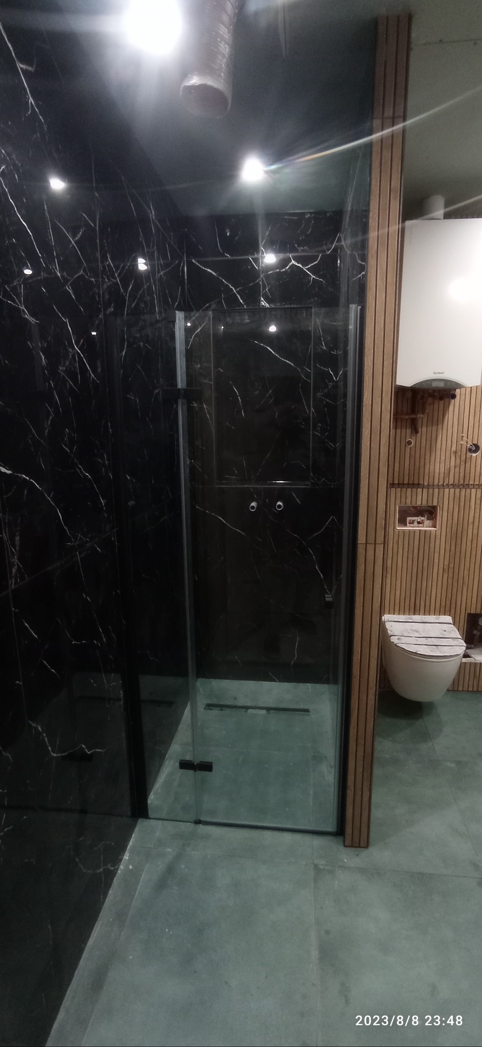 Nowoczesna kabina prysznicowa z czarnymi płytkami imitującymi marmur i szklanymi drzwiami, obok wisząca toaleta i bojler obudowane drewnianymi panelami, oświetlenie punktowe.