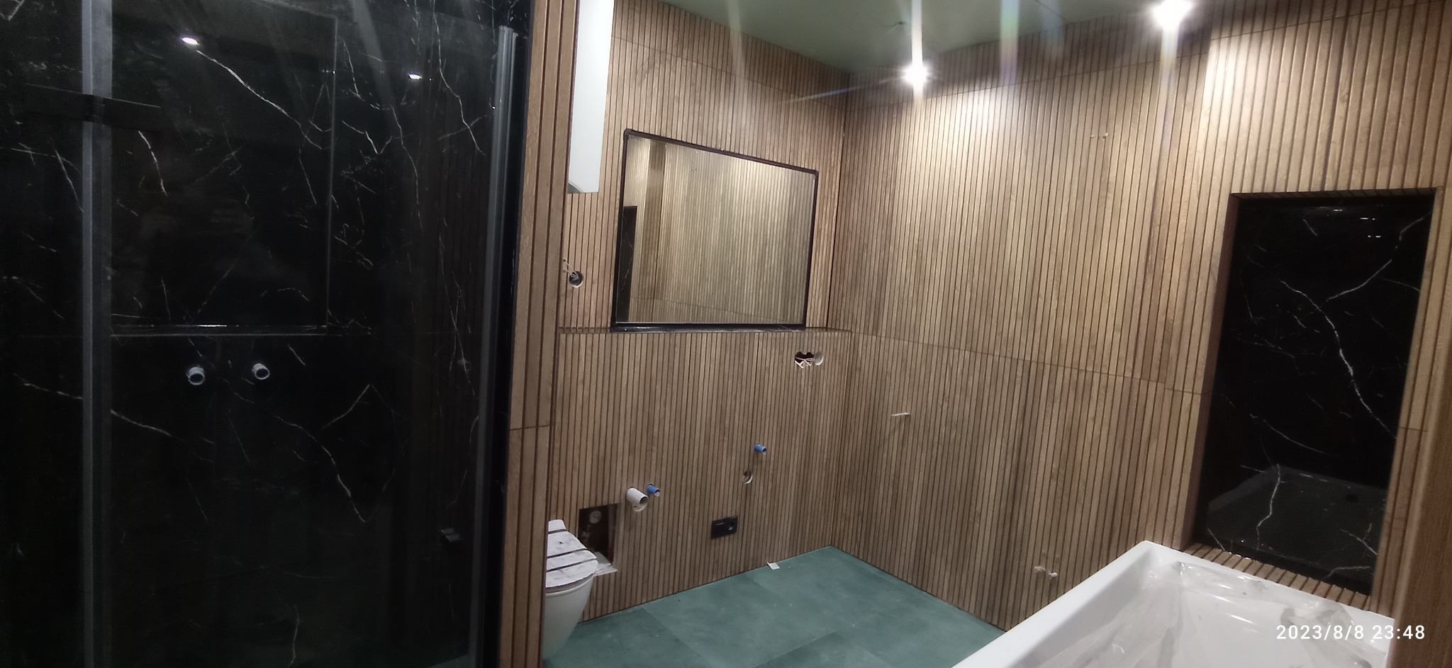 Wnętrze łazienki w trakcie wykończenia: kabina prysznicowa z czarnego marmuru, ściany wyłożone drewnianymi panelami, lustro w czarnej ramie, przygotowane instalacje sanitarne, widoczny fragment wanny.