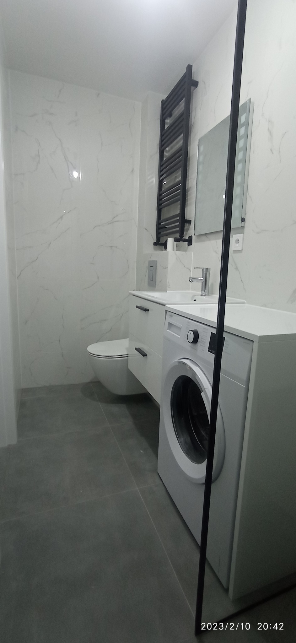 Nowoczesna, jasna łazienka w Oławie po remoncie, z białą armaturą, wiszącą toaletą, umywalką z szafką, pralką i grzejnikiem drabinkowym na ścianie z marmurowym wzorem.