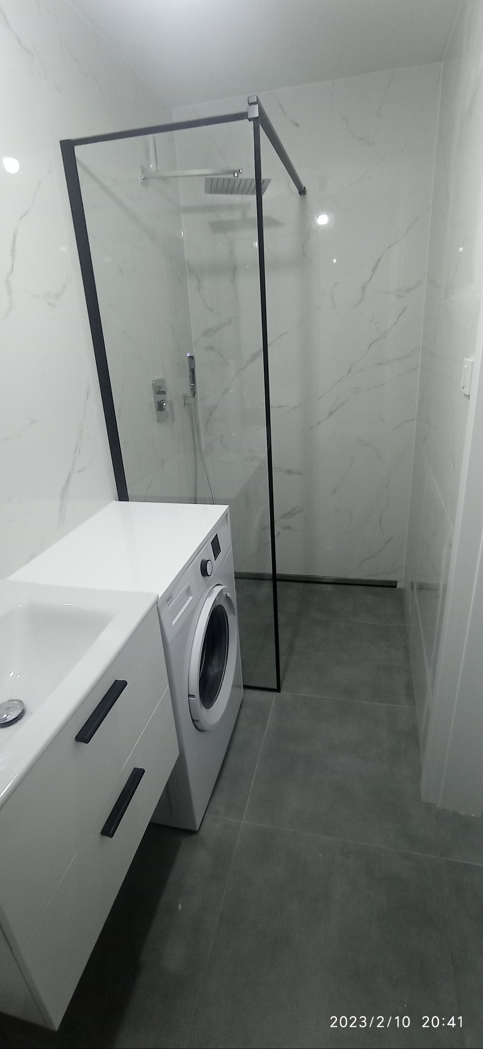 Wykończona łazienka z kabiną prysznicową typu walk-in z czarną ramą, umywalką z szafką z ciemnymi uchwytami i pralką obok, całość w szaro-białej kolorystyce.