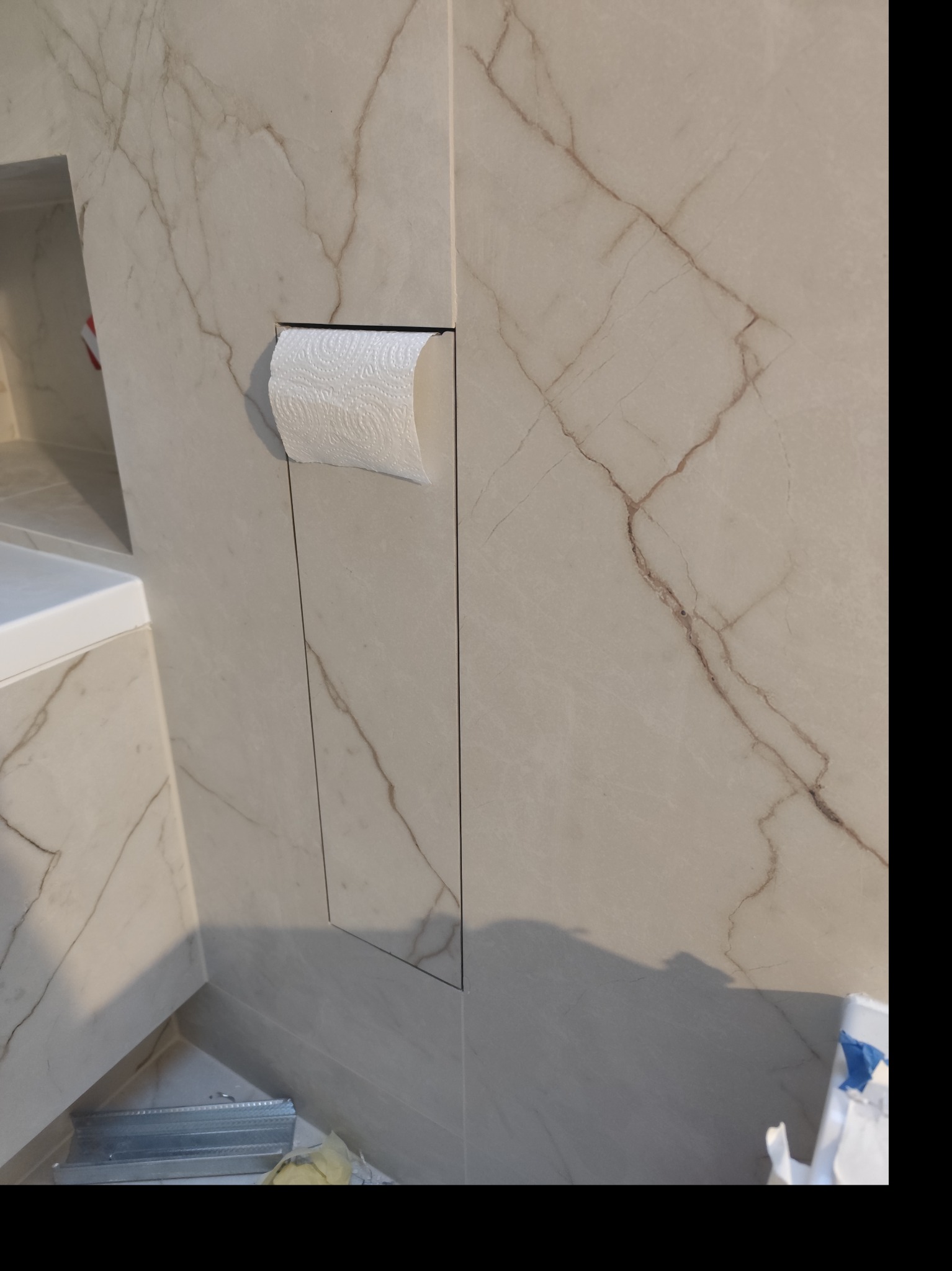 Wykończona łazienka z beżowymi płytkami imitującymi marmur. W ścianie wnęka z ukrytym pojemnikiem na papier toaletowy. Widoczne detale wykończenia i elementy armatury.