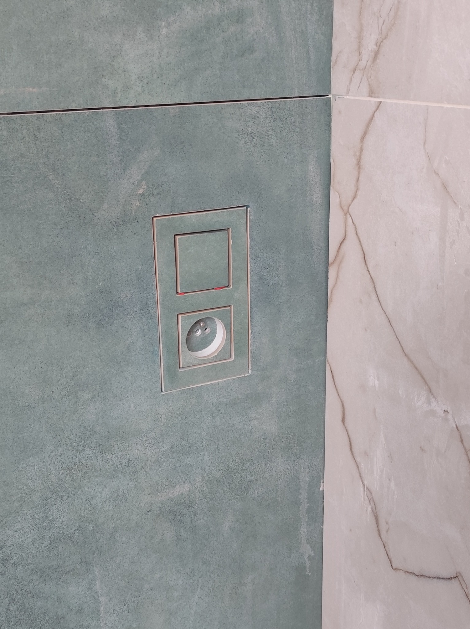 Wykończona ściana w łazience z zielonych płytek z zamontowanym panelem sterującym spłuczki WC. Obok widoczna marmurowa płytka.