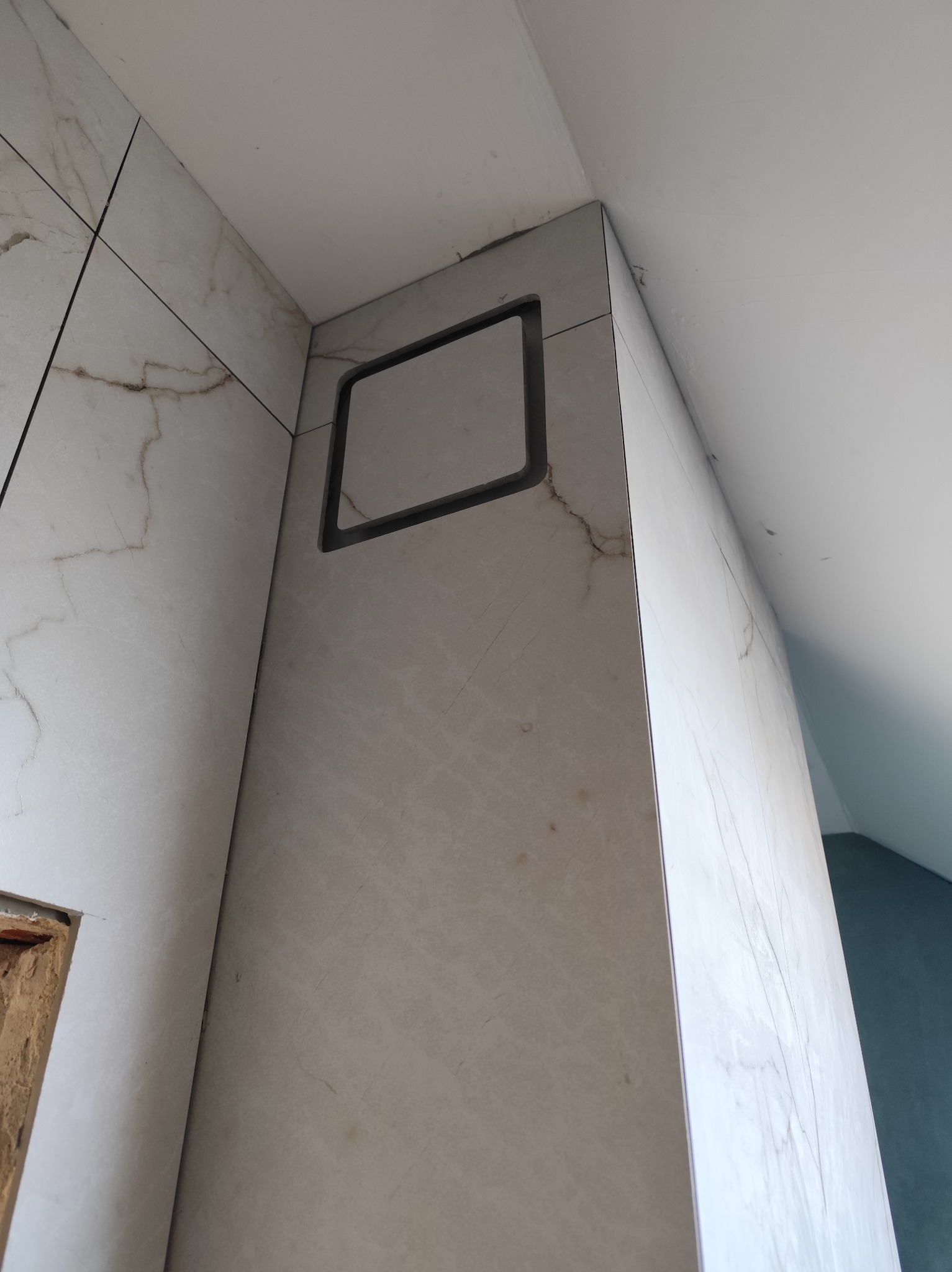 Wykończona ściana w łazience z marmurowymi płytkami i otworem rewizyjnym z czarną ramką. Ujęcie z dołu, widoczny fragment sufitu i ściany bocznej.