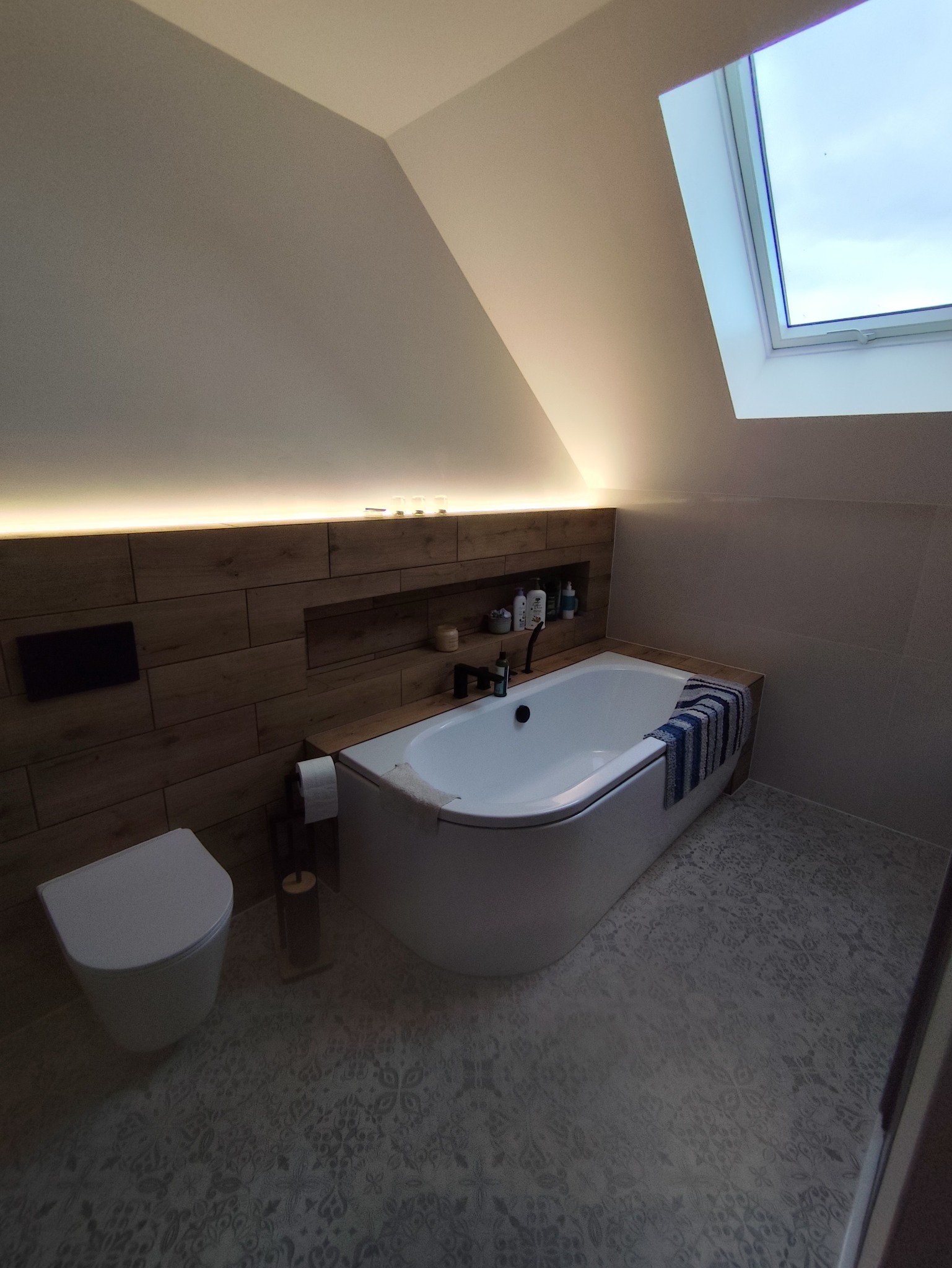Nowoczesna łazienka na poddaszu z wanną, toaletą i drewnianą ścianą z wnęką, oświetlona ledami. Uwagę przykuwa okno dachowe wpuszczające naturalne światło.