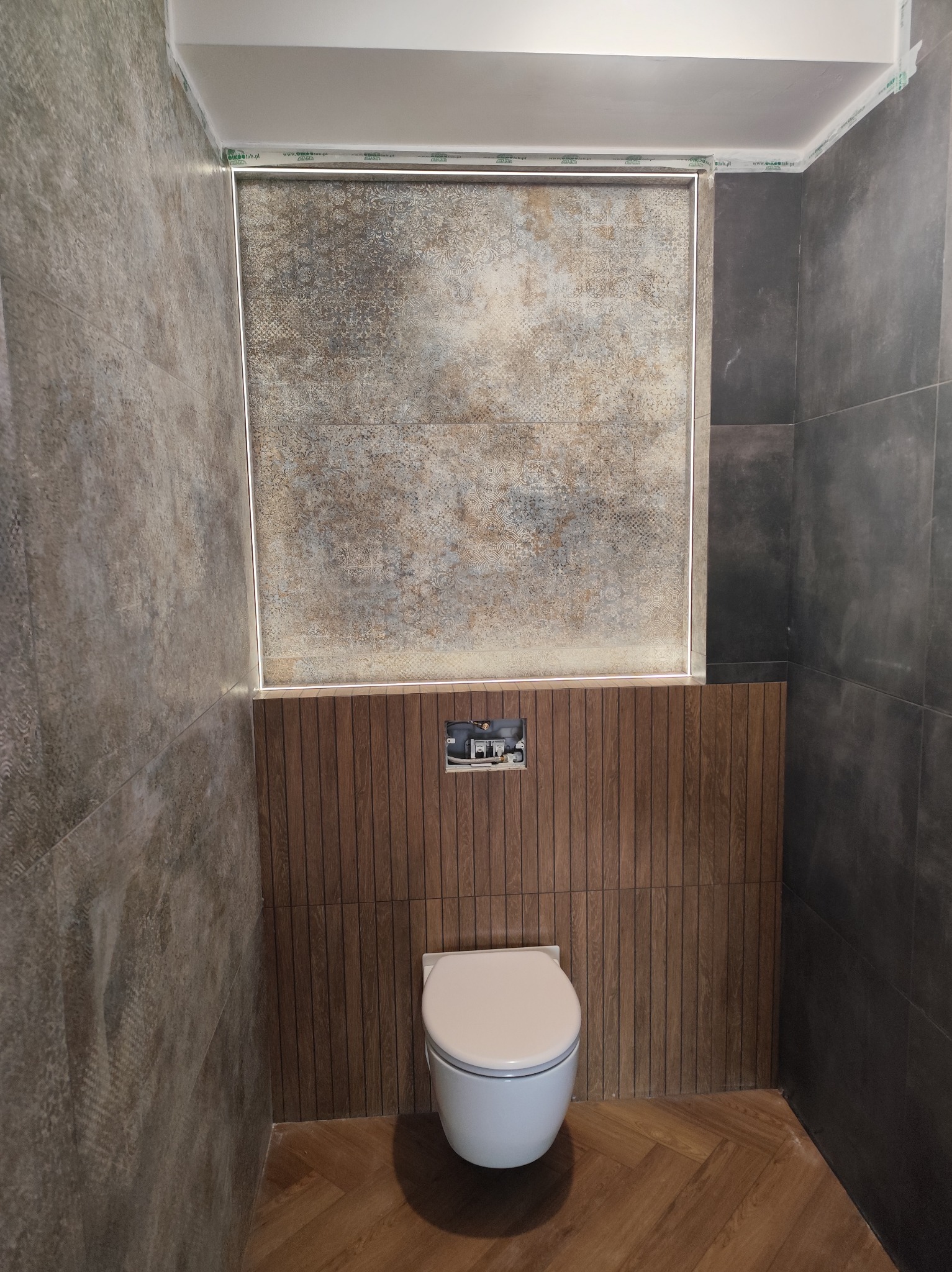 Wykończona łazienka z wiszącą toaletą, brązowymi panelami i szarą mozaiką. Podłoga ułożona w jodełkę. Widoczna instalacja podtynkowa. Minimalistyczny design.