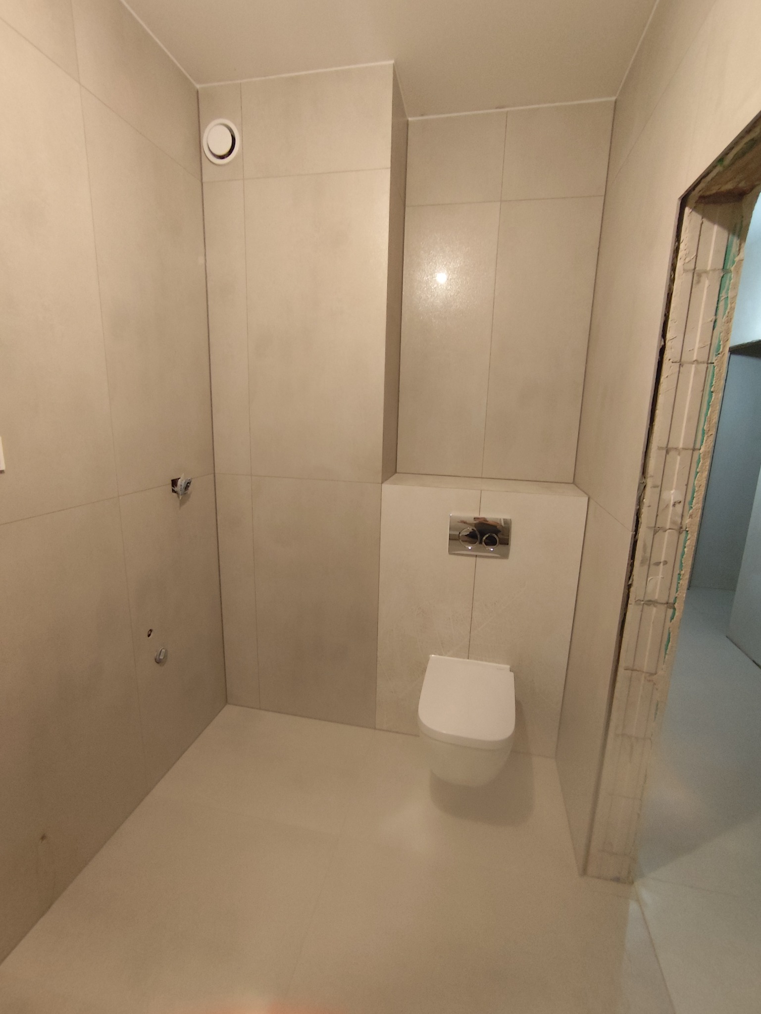 Wykończona łazienka z beżowymi płytkami, wiszącą toaletą i widocznym fragmentem ościeżnicy. Minimalistyczny design, jasne wnętrze.