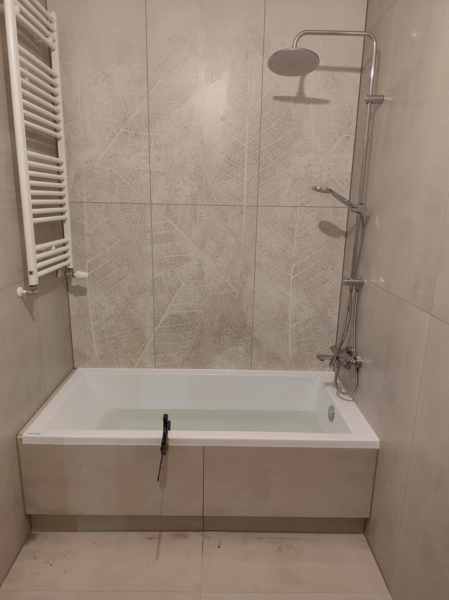 Wykończona łazienka z wanną obudowaną płytkami, grzejnikiem drabinkowym i deszczownicą. Płytki w beżowym odcieniu z delikatnym wzorem liści. Widoczne narzędzie na wannie.