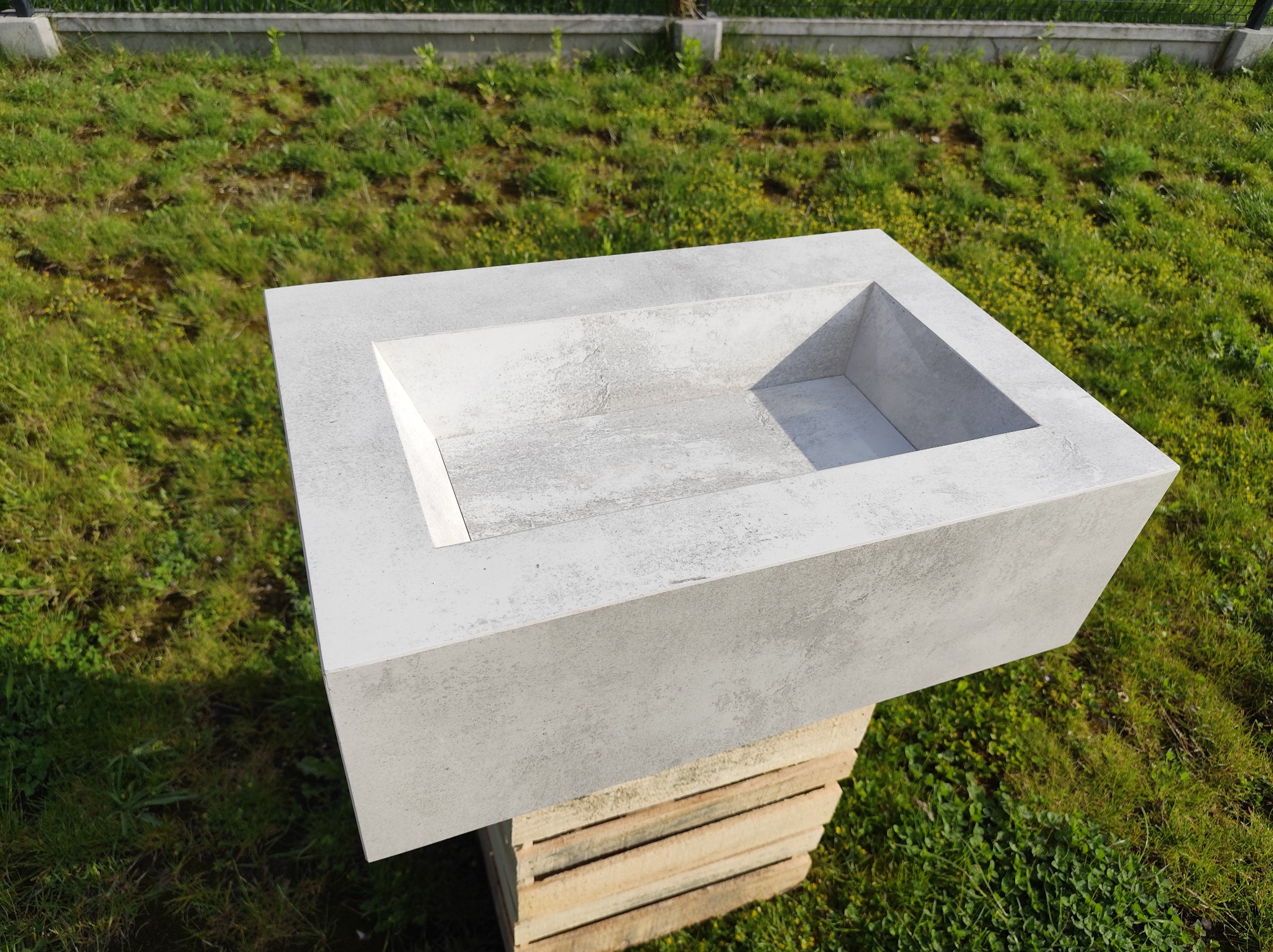 Szara, betonowa umywalka nablatowa o geometrycznym kształcie, ustawiona na drewnianych paletach na tle zielonej trawy. Minimalistyczny design, nowoczesne wykończenie.