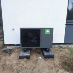 Szara pompa ciepła marki Mycond, zamontowana na betonowych blokach przed biało-szarą ścianą budynku, widoczna butelka wody i samochód w tle.