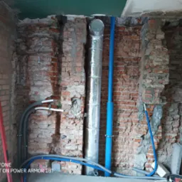 Instalacja rur wentylacyjnych i hydraulicznych w surowym murze z cegły, widoczne rury w izolacji, niebieskie rury PCV i szare rury instalacyjne, poziomica przy ścianie.
