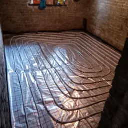 Rury ogrzewania podłogowego ułożone na folii z siatką w pomieszczeniu z ceglanymi ścianami, widok z góry w trakcie montażu.