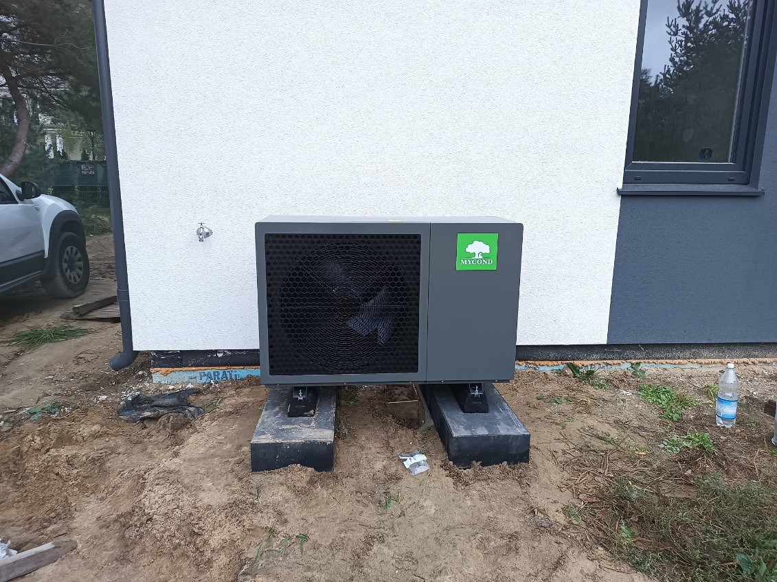 Szara pompa ciepła marki Mycond, zamontowana na betonowych blokach przed biało-szarą ścianą budynku, widoczna butelka wody i samochód w tle.
