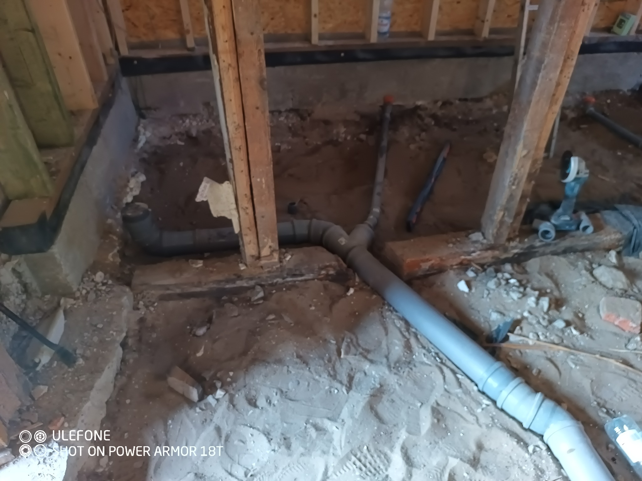 Szara rura kanalizacyjna w trakcie montażu w wykopie ziemnym w pomieszczeniu w trakcie budowy, widoczne drewniane elementy konstrukcyjne i narzędzia.