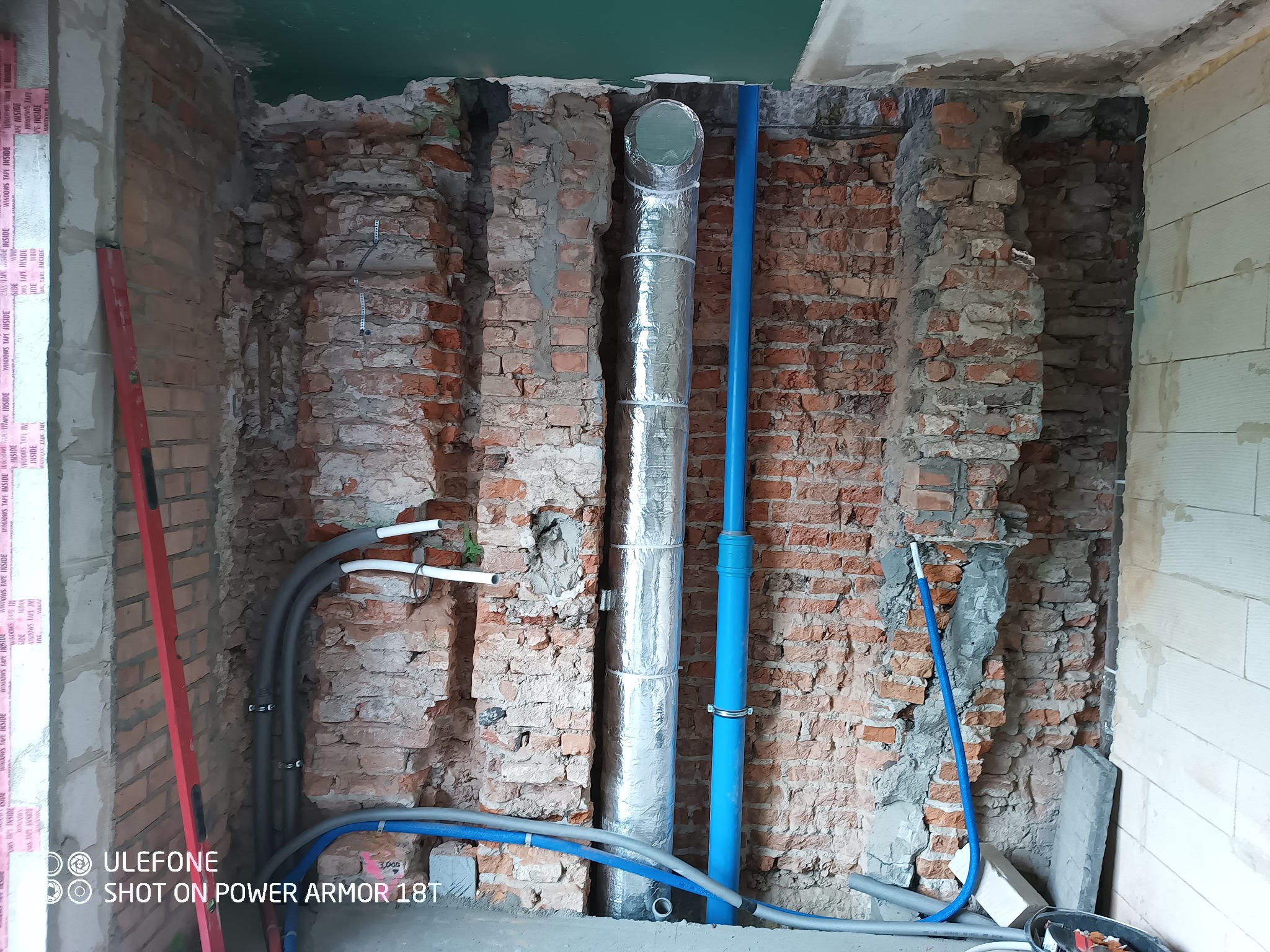 Instalacja rur wentylacyjnych i hydraulicznych w surowym murze z cegły, widoczne rury w izolacji, niebieskie rury PCV i szare rury instalacyjne, poziomica przy ścianie.