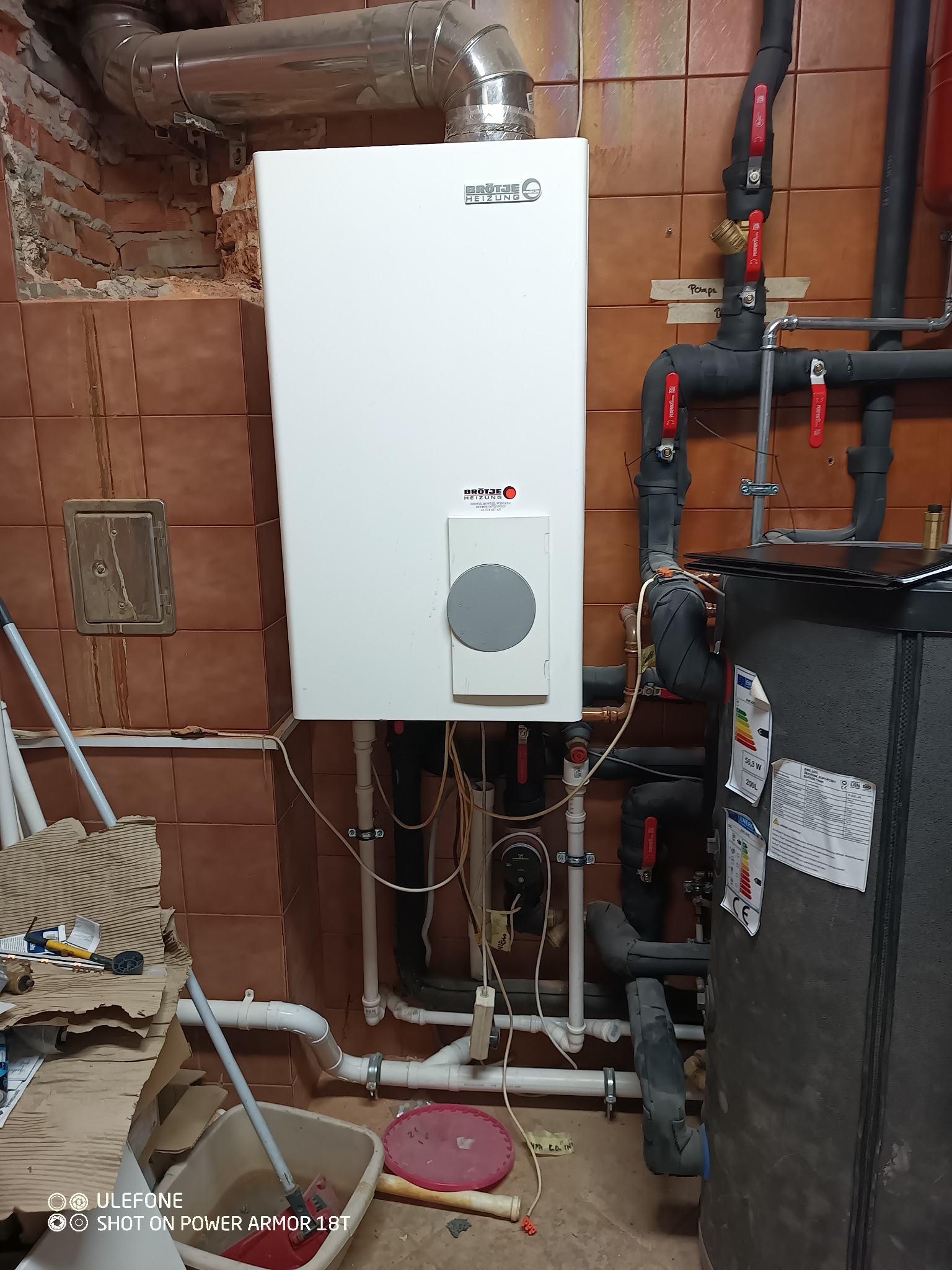 Biały piec gazowy Brotje zamontowany na brązowej ścianie z kafelek, widoczne rury, zawory i zasobnik ciepłej wody, bałagan na podłodze.