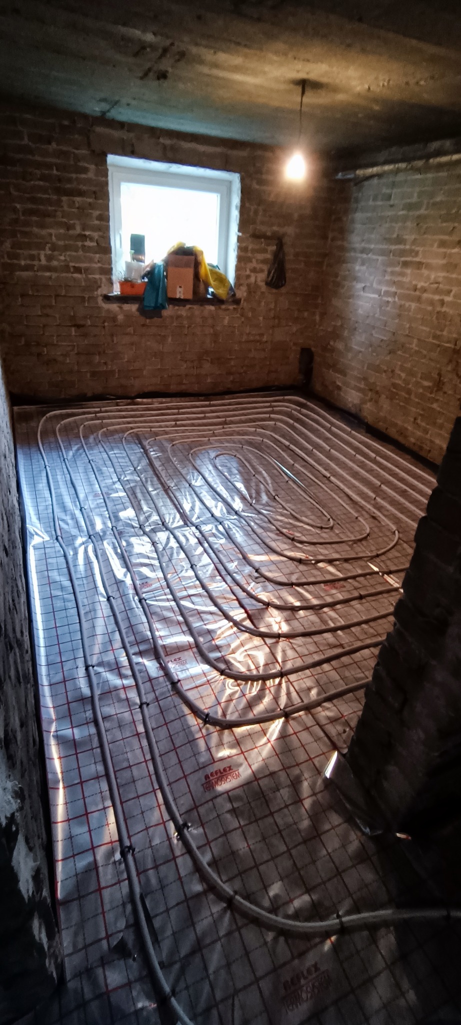 Rury ogrzewania podłogowego ułożone na folii z siatką w pomieszczeniu z ceglanymi ścianami, widok z góry w trakcie montażu.