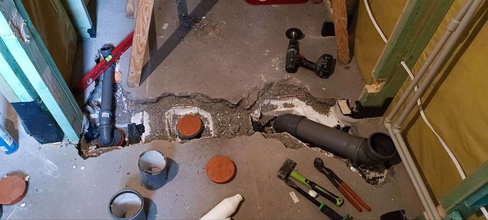 Wykopany betonowy fundament z widocznymi rurami kanalizacyjnymi, narzędziami hydraulicznymi i wiertarką udarową, w trakcie instalacji.