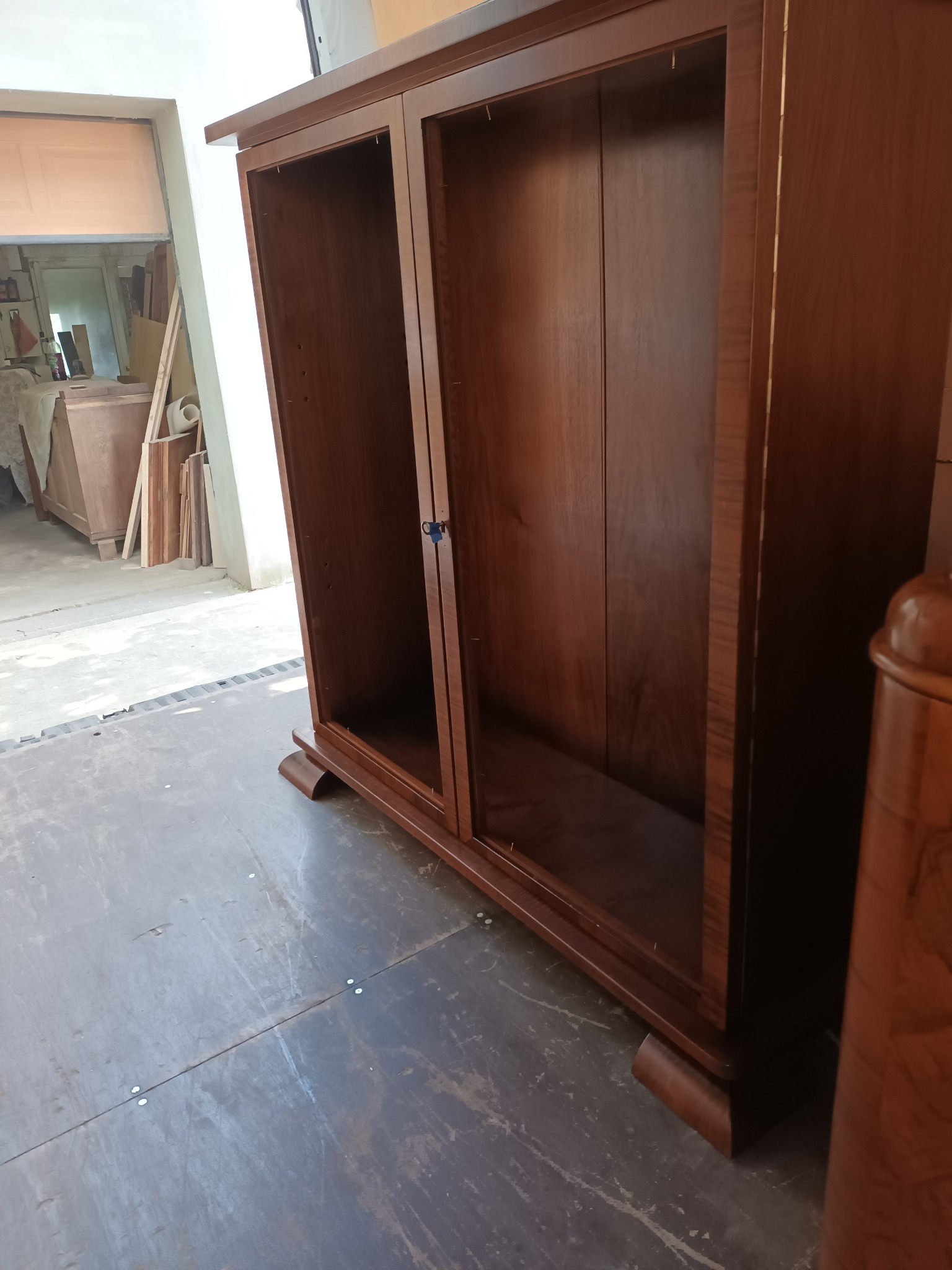 Brązowa, dwudrzwiowa, pusta szafa z drewnianymi nogami, sfotografowana w warsztacie stolarskim, z widocznymi elementami konstrukcyjnymi i wykończeniowymi.