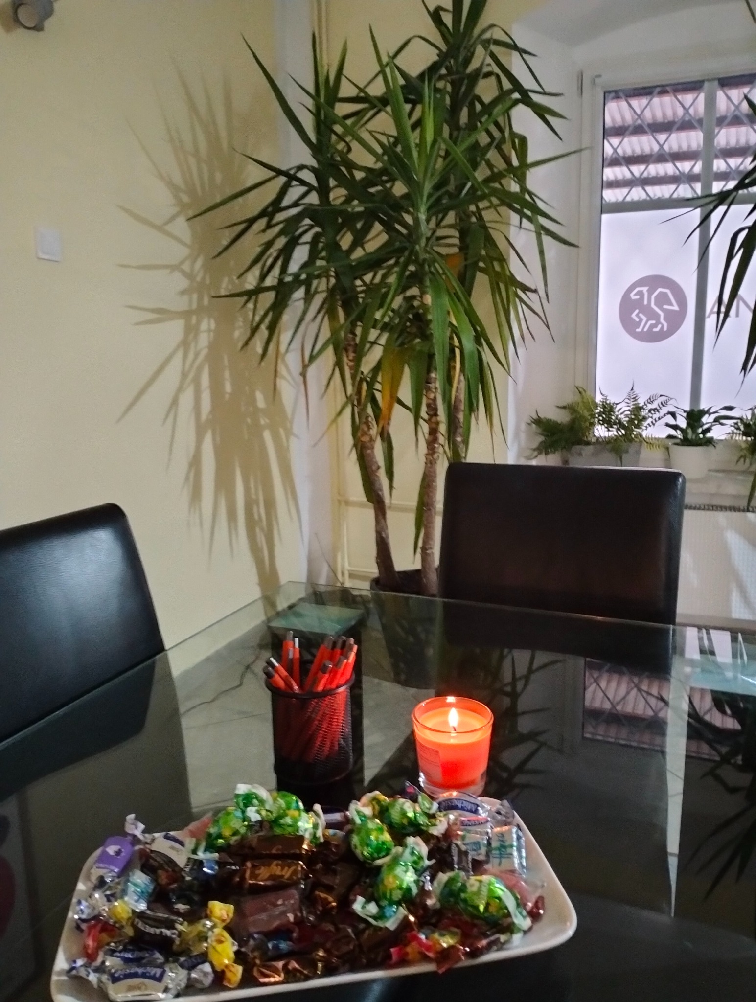 Stół z czarnym blatem, na nim taca ze słodyczami, świeca i pojemnik z długopisami. W tle roślina doniczkowa i okno z logotypem firmy.
