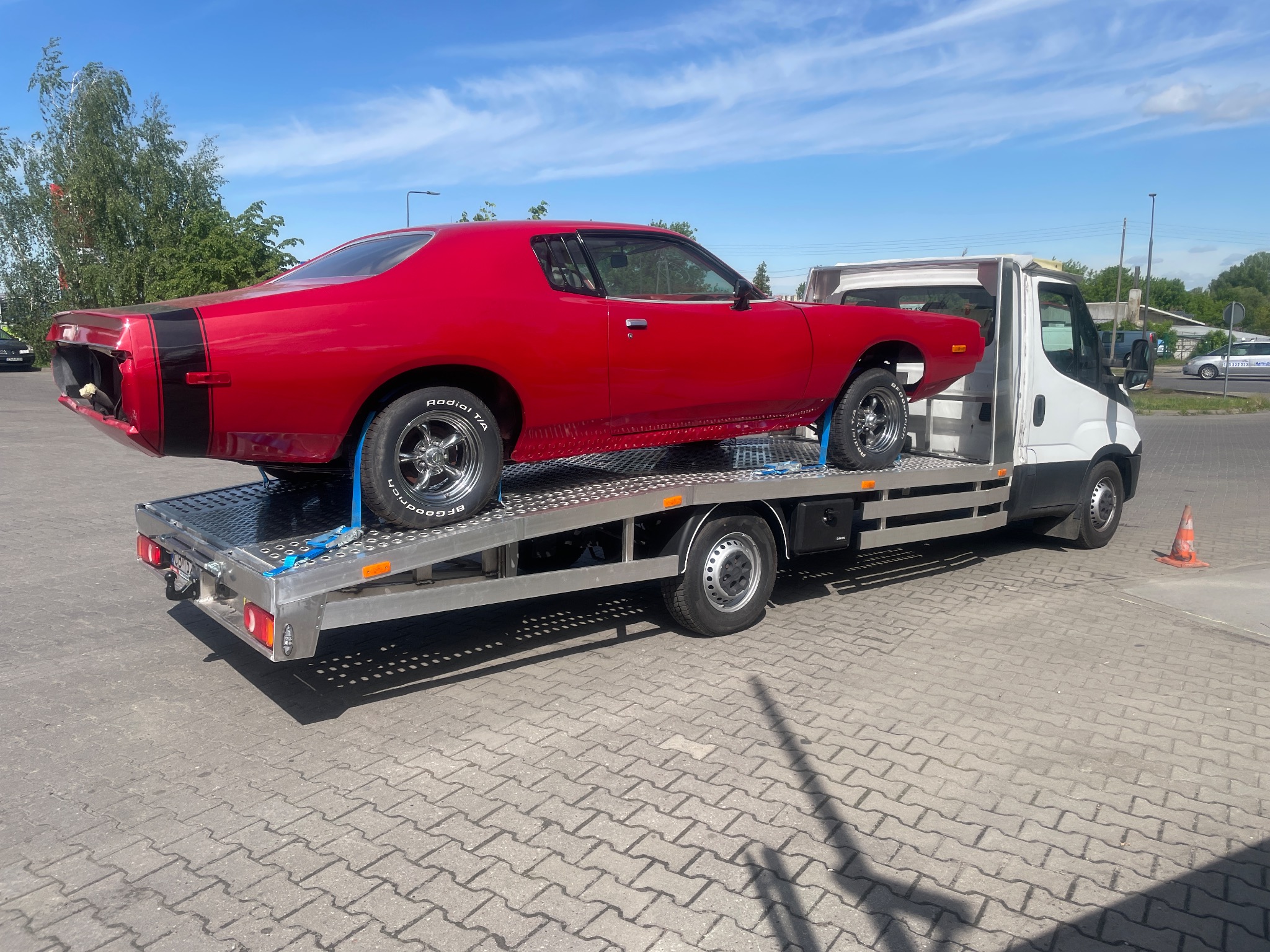Czerwony, klasyczny Dodge Charger z czarnym pasem na tyle, zabezpieczony niebieskimi pasami, transportowany na lawecie w słoneczny dzień.