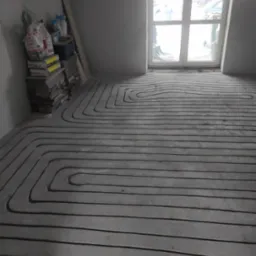 Szare rury w betonie układane w pętle na podłodze w surowym stanie, przygotowane do zalania wylewką, widoczne worki z zaprawą i okno w tle.