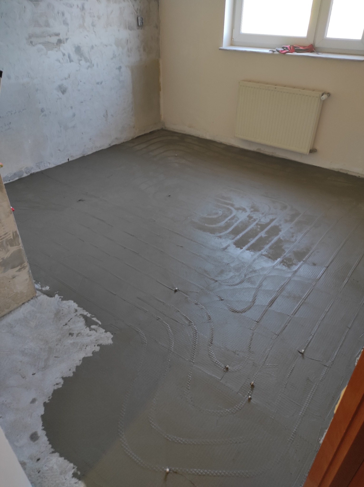 Pokój w trakcie prac wykończeniowych, widoczne zatopione w wylewce rurki instalacji podłogowej, ściany w stanie surowym, grzejnik pod oknem.