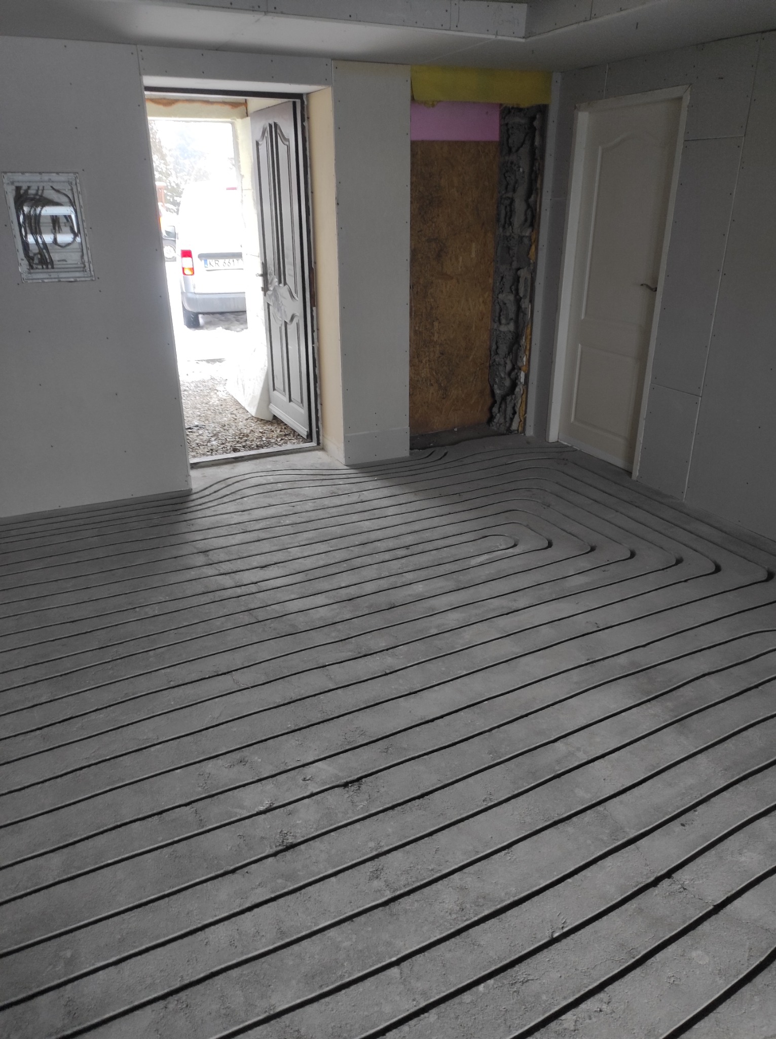 Szare rury ułożone spiralnie w wylewce podłogowej, przygotowane do zalania betonem, widoczne kable w ścianie, otwarte drzwi na zewnątrz.