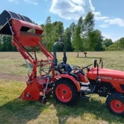 Czerwony traktor Kubota z zamontowanym koszem zbierającym trawę na łące w słoneczny dzień.