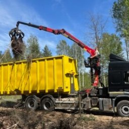 CHOJAK - Czarny samochód ciężarowy z żółtym kontenerem i dźwigiem załadowuje gałęzie drzew. W tle las i fragment domu. Praca w toku.