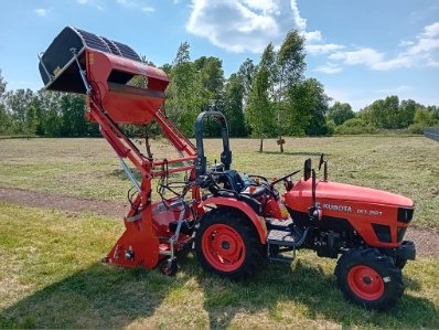 Czerwony traktor Kubota z zamontowanym koszem zbierającym trawę na łące w słoneczny dzień.