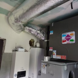 J.K.S company - Szczegółowe ujęcie instalacji wentylacyjnej z widocznym rekuperatorem, kanałami wentylacyjnymi w izolacji aluminiowej oraz dwoma białymi urządzeniami grzewczymi, całość w surowym wnętrzu...